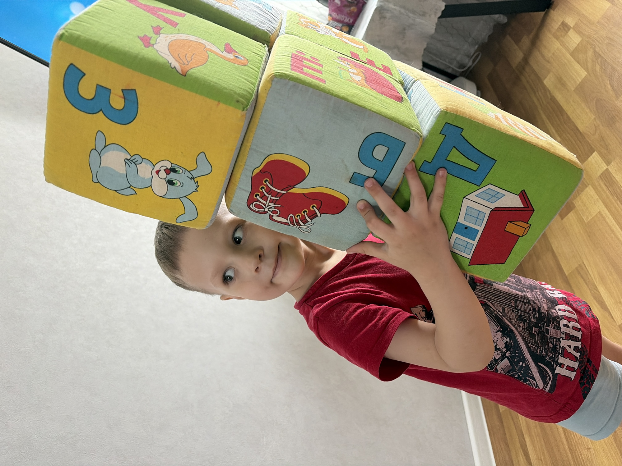 A child wearing a red shirt is holding a stack of soft blocks with images and letters on them. The blocks appear to be alphabet or number blocks designed for toddlers, each having a different color and picture. The child is looking directly at the camera, positioned with their head near the bottom left of the frame. The background consists of a light-colored wall. The environment is indoors with natural light.