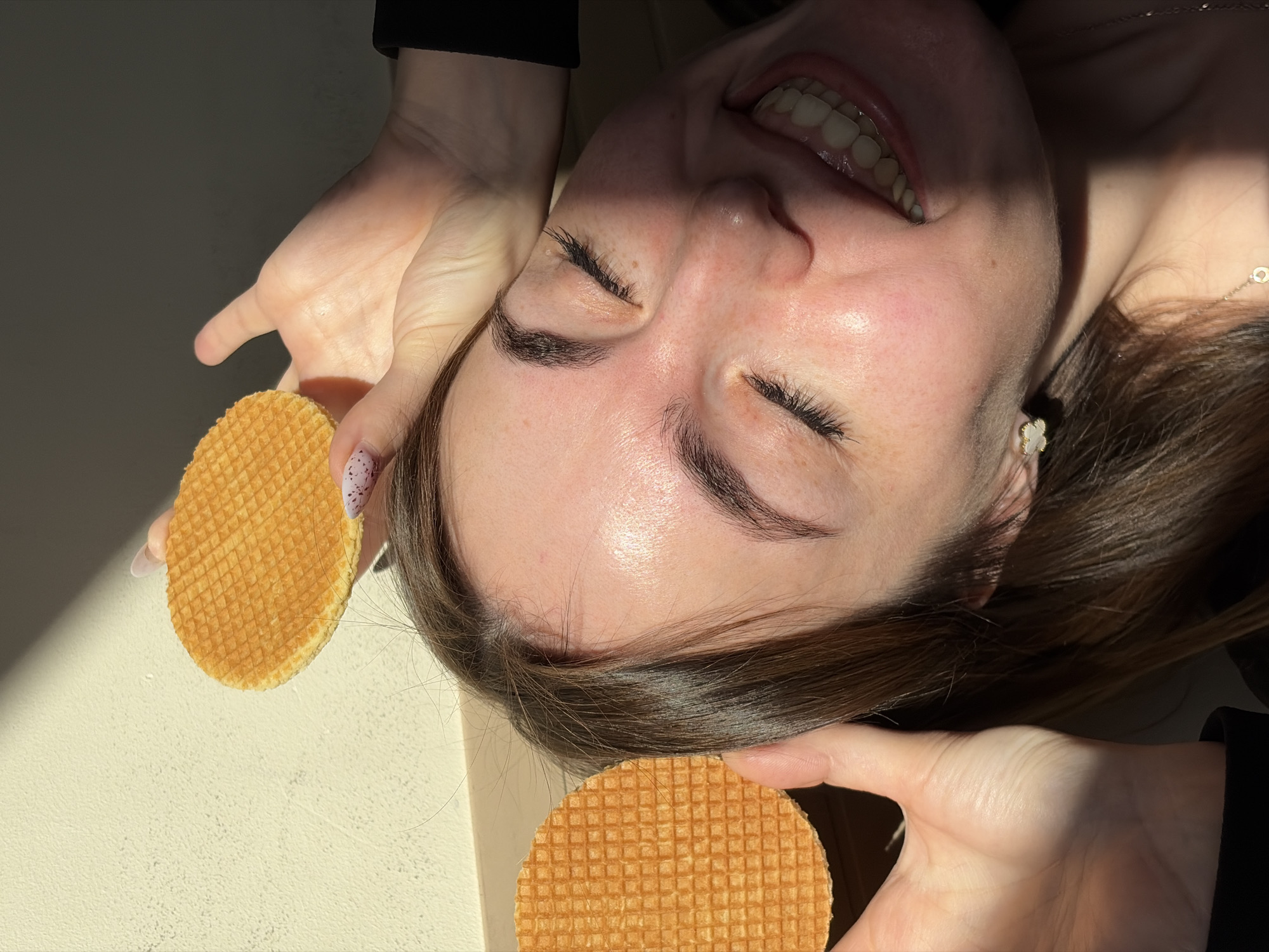 A woman is smiling while lying on a light-colored surface, holding two stroopwafels, one in each hand. The woman has brown hair and her eyes are closed. She is wearing a light-colored earring and a necklace. The background is a light-colored, possibly white, surface, with some shadows. There are no signs of the city Izhevsk, Russia.