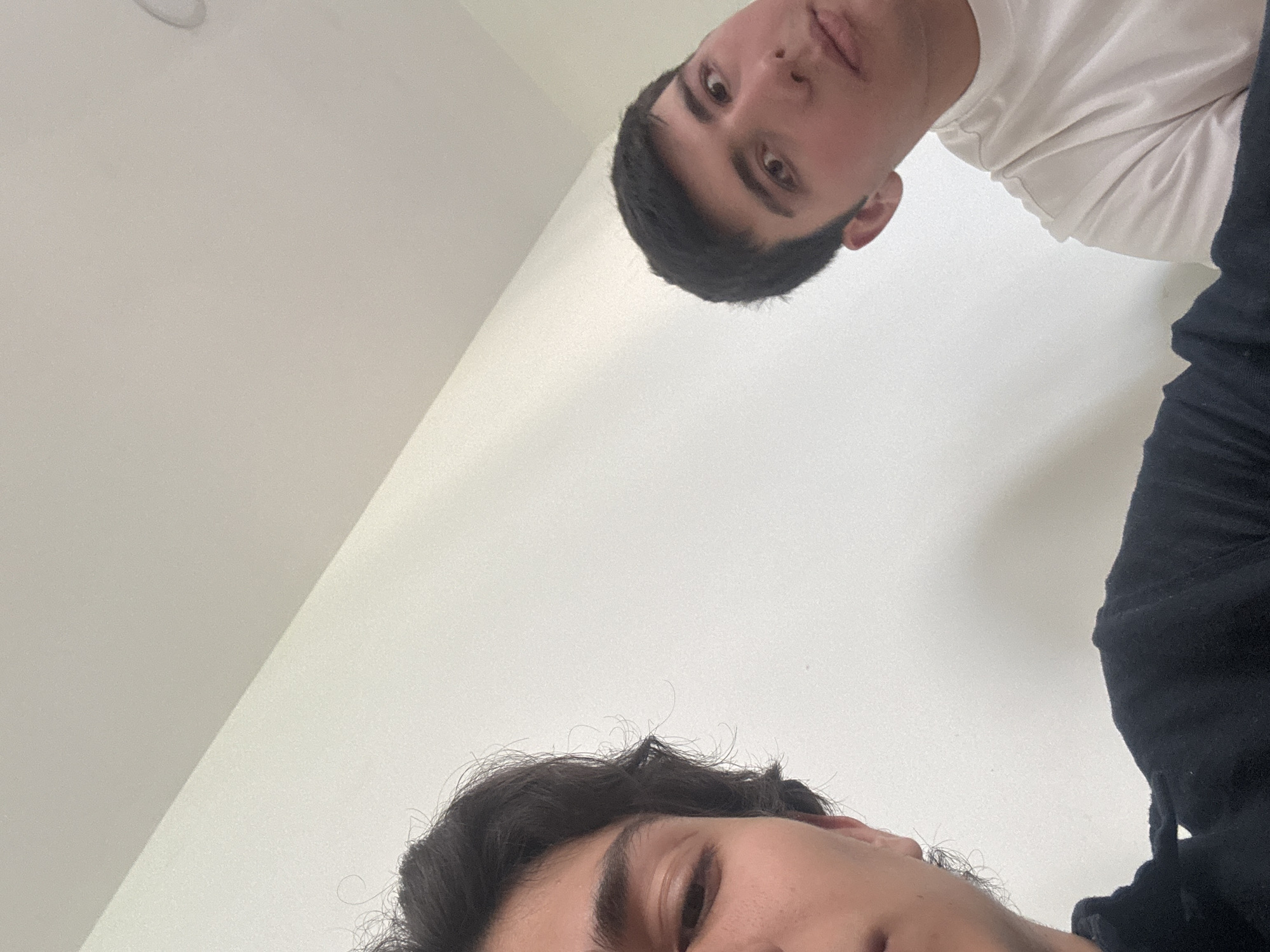 The image shows two people from a low angle perspective, as if the photo was taken from below. One person is wearing a white shirt and has short dark hair. The other person is wearing a black top and has long dark hair. The background is mostly a plain white surface, likely a ceiling or wall. The location is not identifiable from the image content.