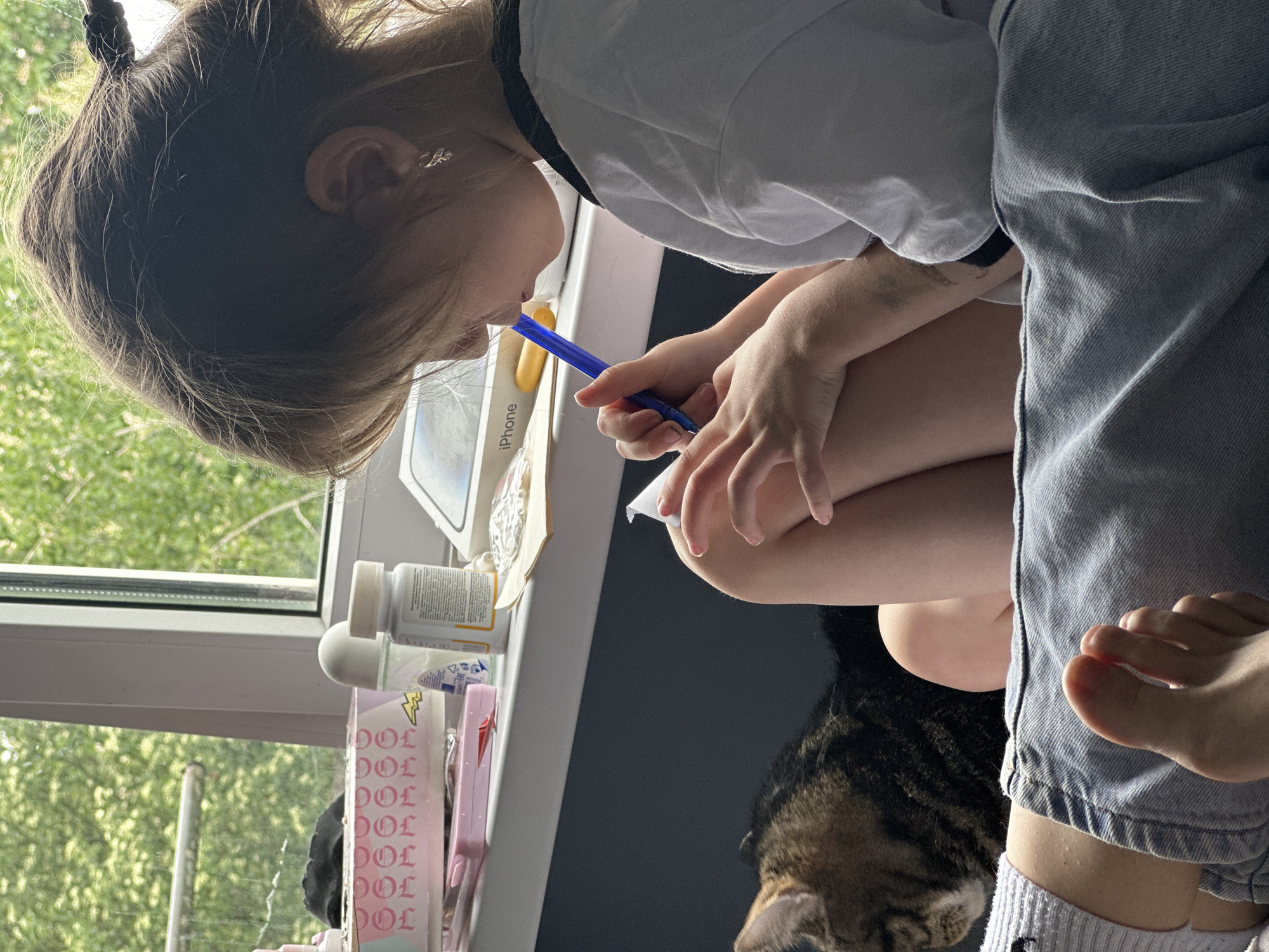 A young person, possibly a child, is seated indoors next to a window with green foliage visible outside. The person is leaning towards a window sill, where various items are placed, including an iPhone box, pill bottles, and other small objects. They are holding a blue pen and a small piece of paper, possibly writing or drawing. A tabby cat is partially visible at the bottom of the image, near the person's legs. The person is wearing light-colored clothing and denim trousers. The location is Stavropol, Russia.