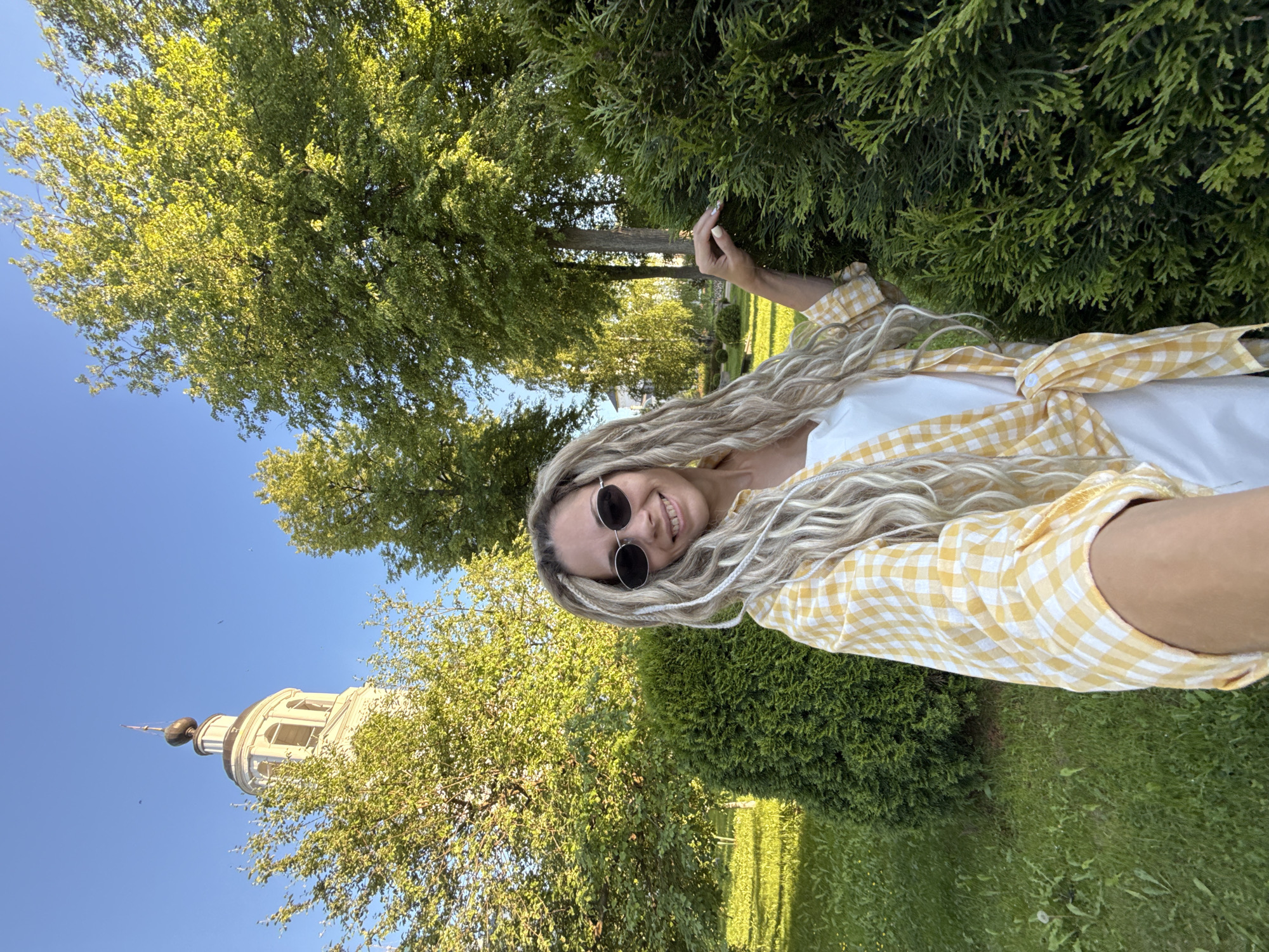 An outdoor, eye-level selfie captures a smiling woman with long, blonde dreadlocks and dark oval sunglasses, looking directly at the camera. She wears a white top under a yellow and white gingham shirt, tied at the waist. Her right hand holds the camera, while her left hand gently rests on a dark green, well-trimmed conifer bush to her right. The scene is set in a sunny park with bright green deciduous trees in the midground and background, and a neatly mowed grassy lawn in the foreground. In the upper left background, partially obscured by tree foliage, stands the light-colored, multi-tiered Bell Tower of the Assumption Cathedral (Uspensky Sobor) in Yaroslavl, Russia, featuring arched belfry openings, a dark onion-shaped cupola, and a slender spire. The sky above is clear and blue.