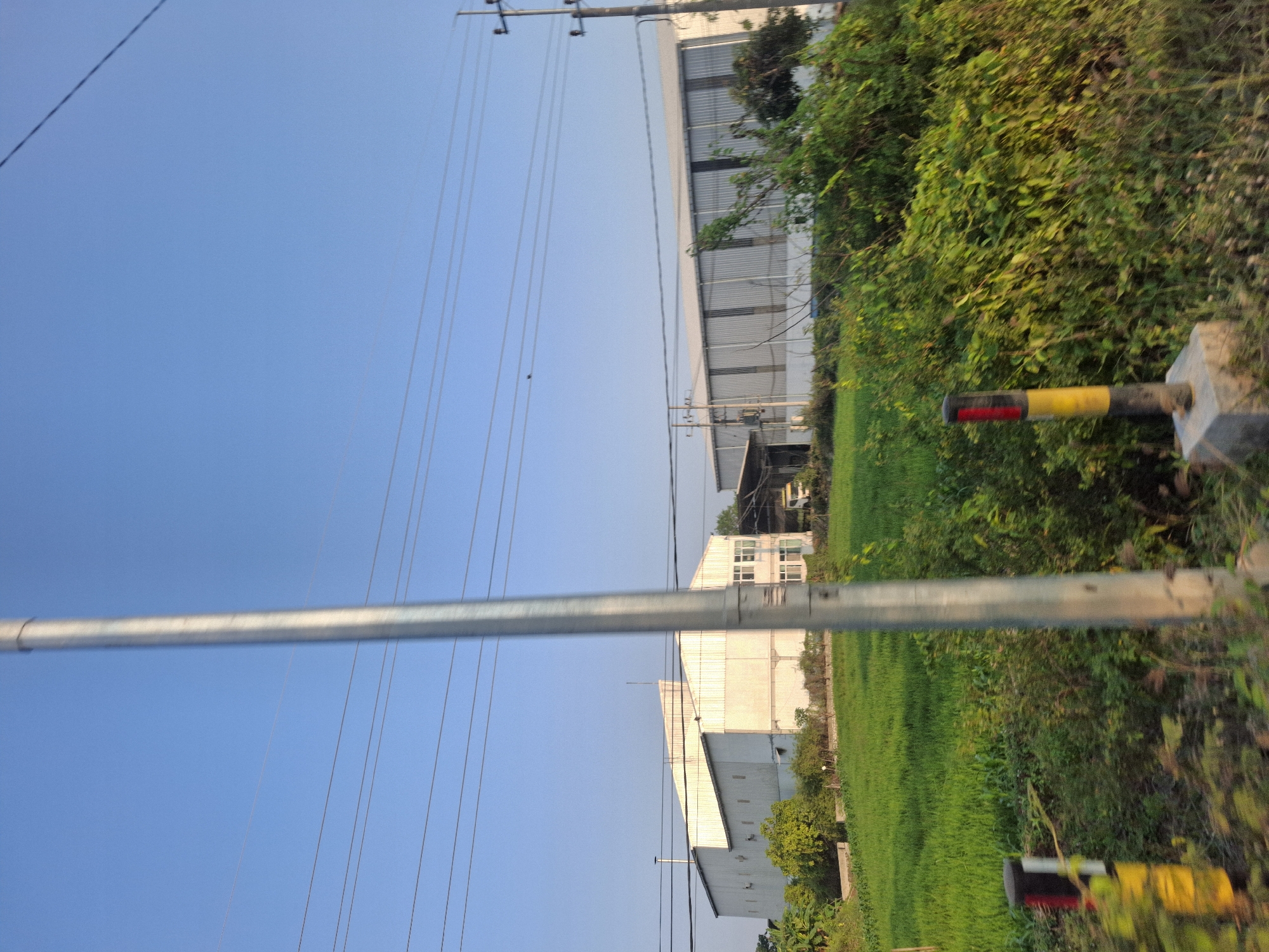 This photograph captures a sunlit scene in the vicinity of Sungelebak, Indonesia, characterized by a clear blue sky and the presence of industrial buildings amidst lush green vegetation. Dominating the foreground is a tall, metallic utility pole, casting a slight shadow. To the left of the pole, a low-lying marker post, painted with black, yellow, and red bands, stands firmly on a concrete base.
The midground reveals two large, white industrial structures. The building on the left, a long, angular structure, features numerous small windows and a distinct chimney. Adjacent to it, to the right, is another elongated building with a corrugated metal facade. A small white truck is visible parked near the entrance of this second building. Between the utility pole and the industrial buildings, a vibrant green field stretches out, likely agricultural land.
The background is a clear, unblemished sky, with several power lines strung across it, supported by poles visible on the right edge of the frame. The overall environment appears to be rural or on the outskirts of a town, given the combination of industrial facilities and expansive green spaces. The lighting suggests it is daytime, with the sun illuminating the scene.