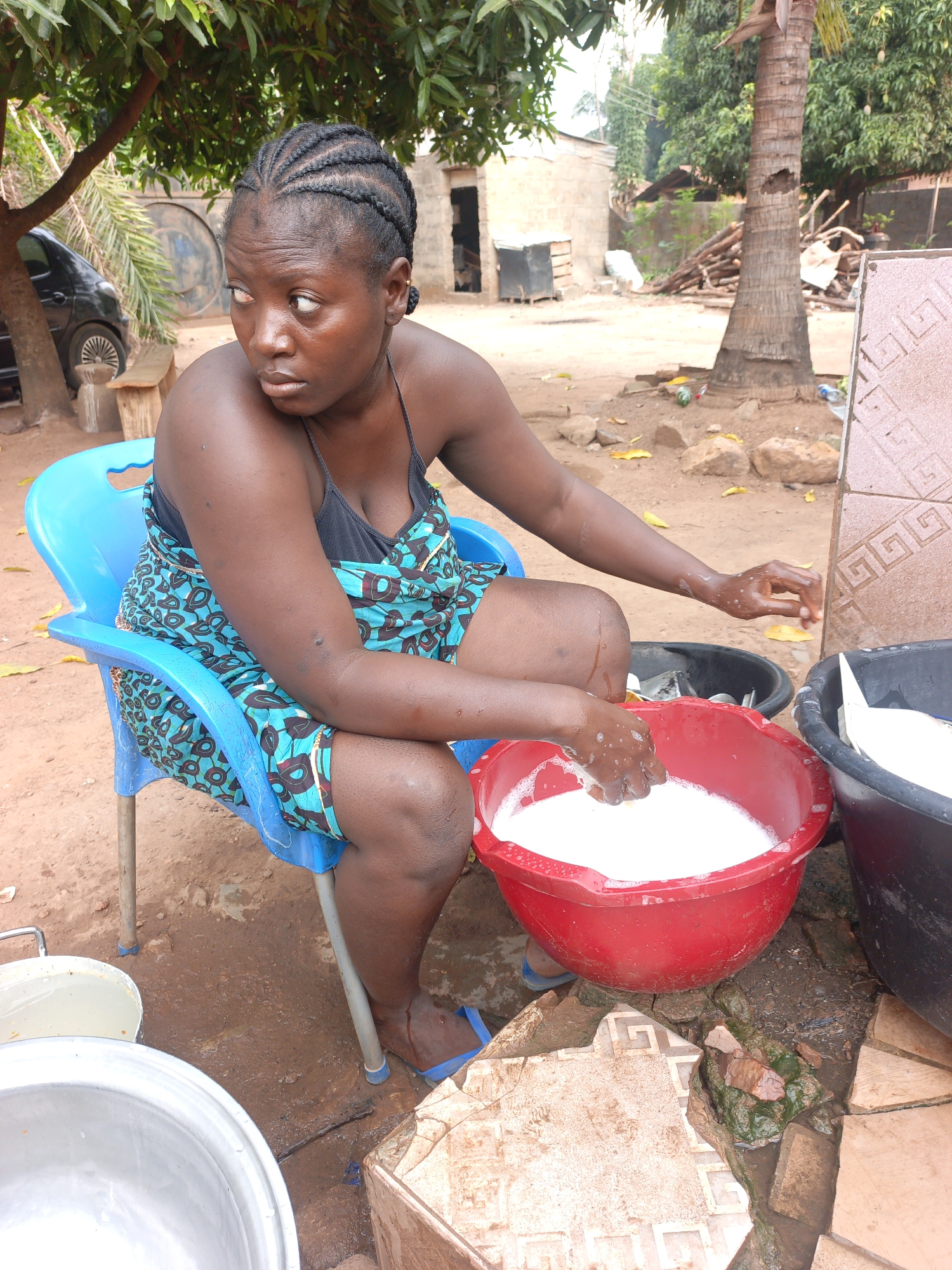 A woman with braided hair sits on a blue plastic chair outdoors, her left arm resting on her thigh and her right hand extended toward a red plastic tub filled with a white, foamy substance. The woman is wearing a black tank top and a blue patterned cloth dress. A black tub sits next to the red one, also filled with a white substance. A silver metal basin is visible in the foreground. The ground is dirt and broken tiles. A building and palm tree are visible in the background. The setting is a yard in Akwanga, Nigeria.