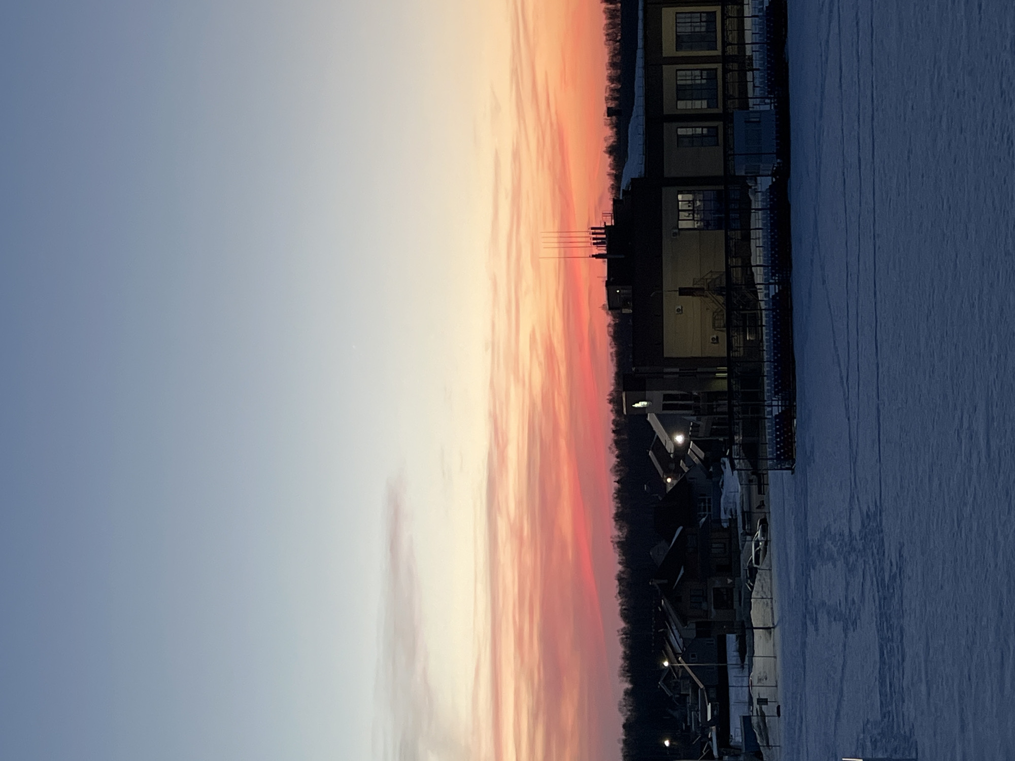 The image presents a panoramic view of Noviy, Russia, during low light conditions, indicative of either dusk or dawn, in a winter setting. The sky transitions from a pale blue at the top to vibrant horizontal streaks of pink and orange clouds across the mid-section, darkening to a deep orange-red closer to the horizon. A faint crescent moon is visible in the upper left quadrant of the sky.
A dense line of dark, leafless trees forms the primary horizon line. Below the tree line, a populated area is visible. On the left, a cluster of residential houses with dark roofs and several illuminated windows are present. Streetlights are active, casting light onto snow-covered surfaces. Two vehicles are discernible parked on a snow patch near these houses. On the right, a multi-story commercial or public building with a flat roof and multiple illuminated windows stands prominently. Its roof features several vertical spires or antennas. Adjacent to this building are rows of exterior seating, resembling bleachers, and a small, light-colored, enclosed structure.
The foreground is dominated by a large, flat expanse of snow, upon which numerous faint tracks and lines are visible. A partial white vertical pole or marker is present in the far left foreground. No individuals are clearly depicted in the scene. The entire ground is covered in snow.