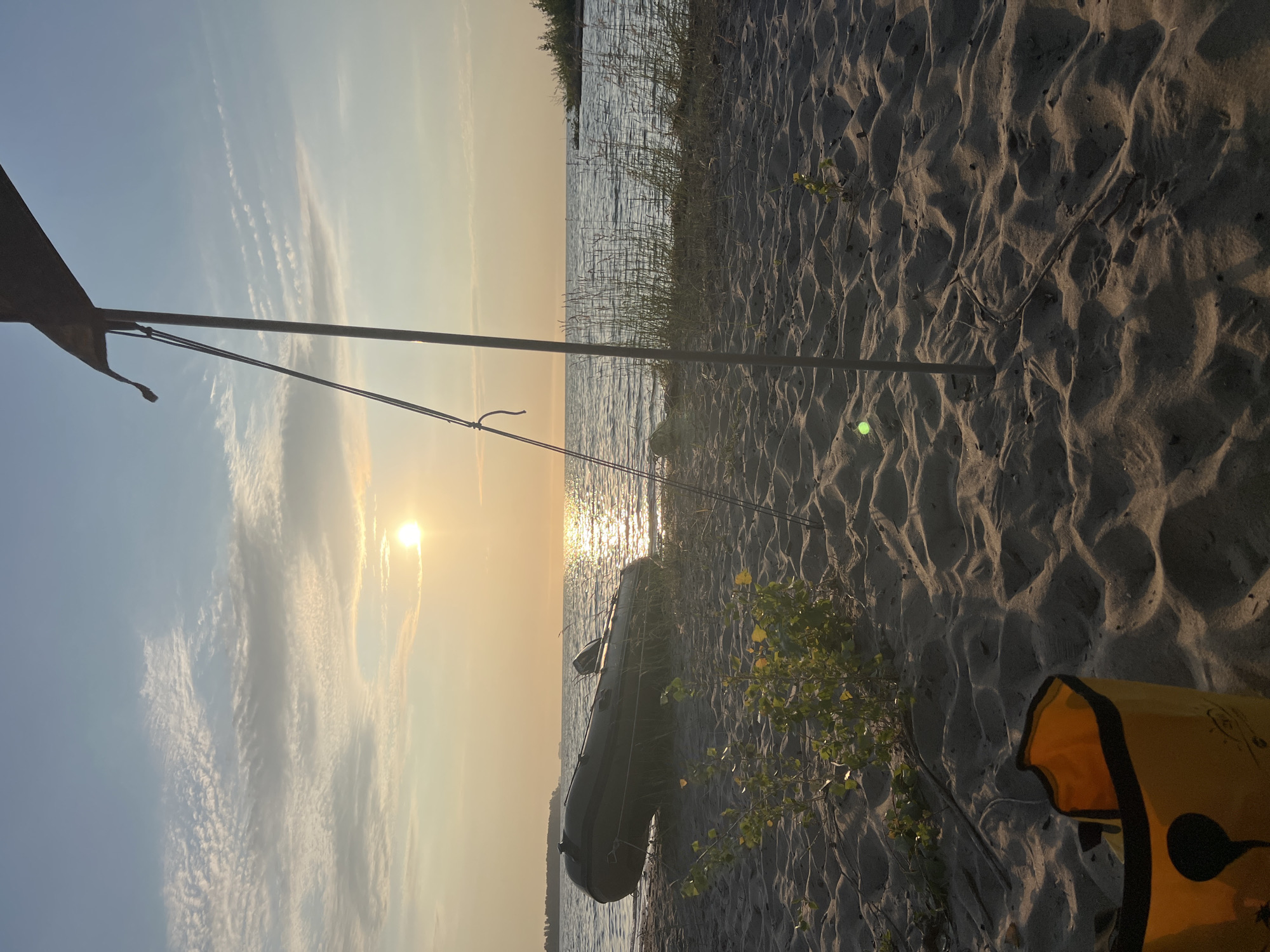 The image depicts a serene beach scene at sunset, likely in Borovoye, Russia. The sun, a bright orb, is low on the horizon, casting a warm, golden glow across the sky and water. Wispy clouds are scattered across the pale blue sky. In the foreground, sand dunes are textured with footprints and scattered with small twigs and patches of green vegetation. A yellow bag with black trim is visible in the lower left corner. To the left, a dark green inflatable boat rests on the sandy shore, its motor clearly visible. A tall, thin metal pole, part of what appears to be a canopy or tent structure, stands in the middle ground, with ropes extending from it. Tall grasses grow along the edge of the water. The calm, shimmering water of a lake or sea stretches to the horizon, with a distant tree-lined shore visible on the right. The overall atmosphere is tranquil and suggestive of a peaceful evening spent outdoors.