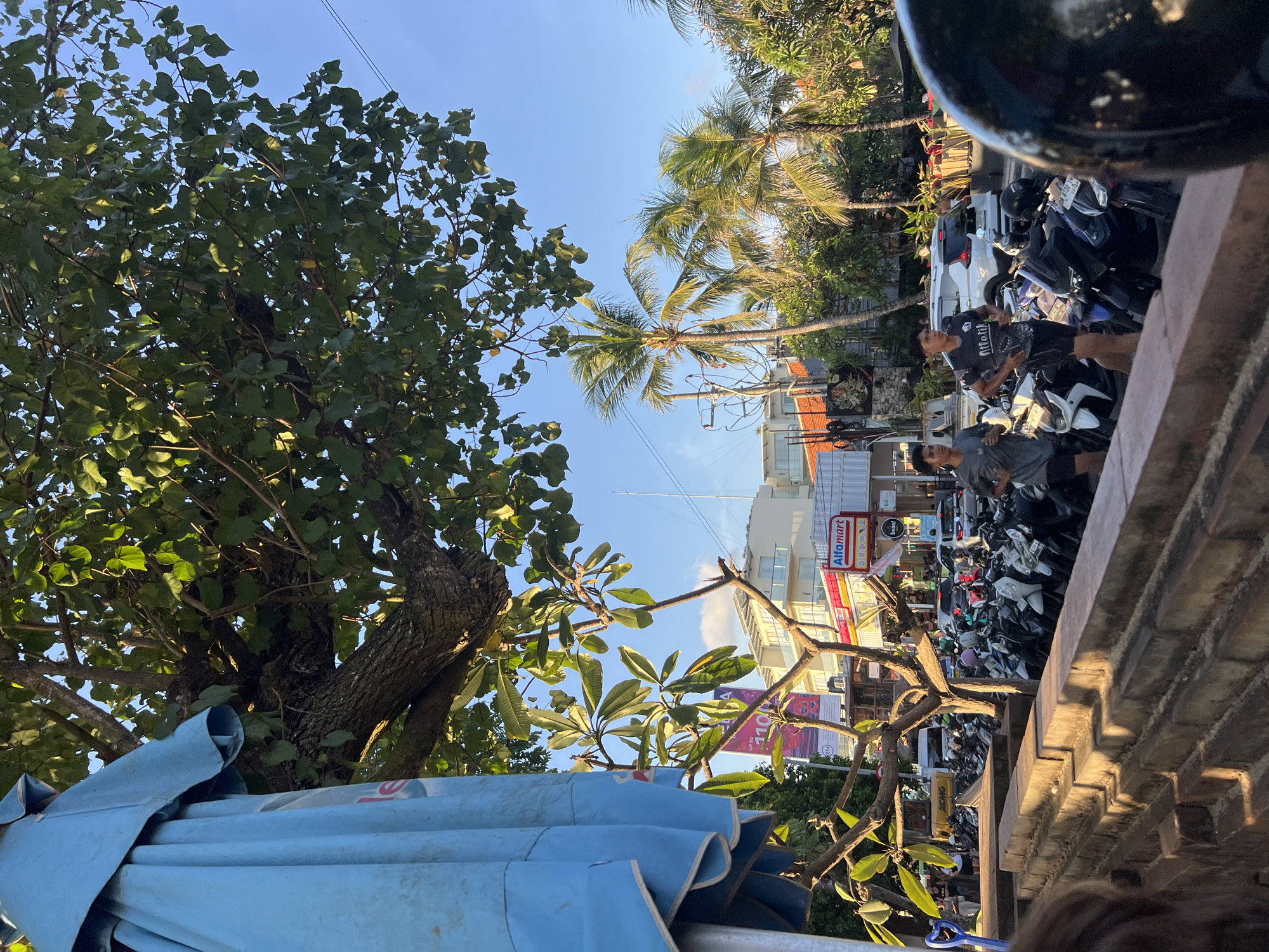 In Kuta, Indonesia, a street scene unfolds under a clear blue sky, bathed in the warm glow of late afternoon sunlight. A large blue umbrella, partially visible on the left, casts a shadow over the foreground, where a weathered brick wall forms a textured ledge. Beyond the wall, a row of parked motorcycles lines the street, their varied colors and designs creating a mosaic of urban transportation.
Two young men are captured mid-stride, jogging along the sidewalk. The man on the left wears a grey t-shirt and black shorts, while the man on the right sports a dark t-shirt with a graphic design and black shorts. They appear to be running in the same direction, their bodies angled towards the right side of the frame.
In the background, palm trees sway gently, their fronds silhouetted against the bright sky. Buildings with white facades and balconies are visible behind the trees, suggesting a commercial or residential area. A sign for "Alfamart," a popular convenience store chain in Indonesia, is prominently displayed on one of the buildings, indicating a local commercial hub. The overall atmosphere is one of a vibrant street scene, likely a popular thoroughfare in Kuta, with a mix of natural elements and urban activity.