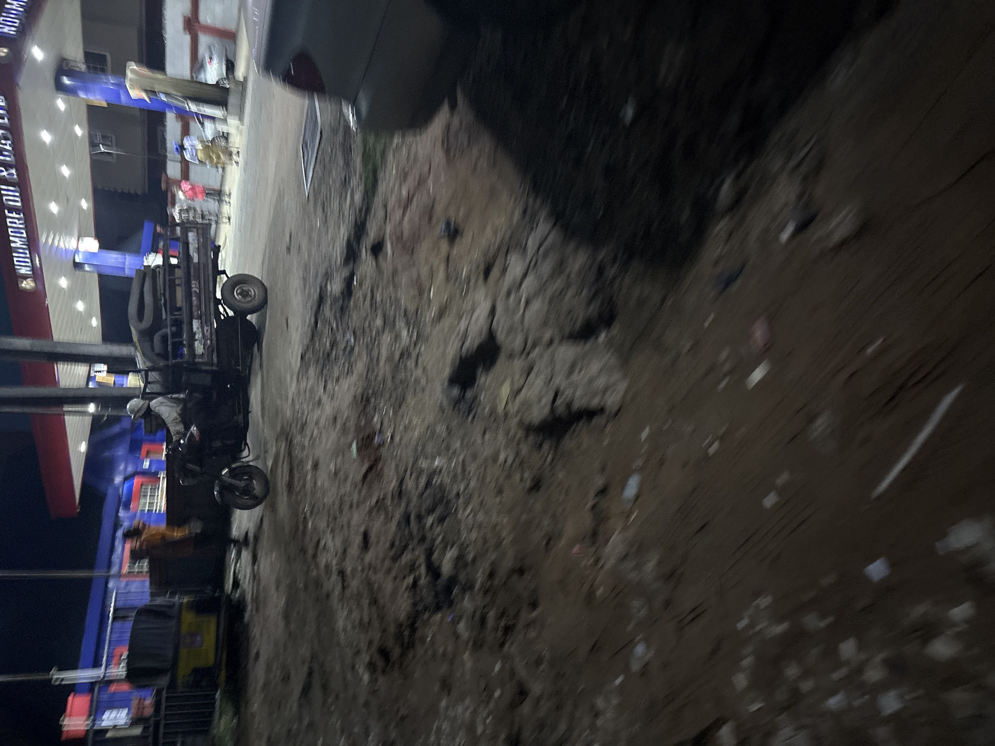 An outdoor, night scene at a Nolmore Oil & Gas Ltd. filling station in Nkpor, Nigeria shows a person driving a motorized tricycle with a flatbed carrying rolled-up foam. The tricycle is positioned in front of the gas station canopy. To the left, another motorized tricycle, yellow and black, is parked. A person in an orange shirt stands near it. In the background, people are visible near the gas pumps, and a silver car is parked to the right. The ground in the foreground and mid-ground is muddy and uneven, with scattered debris. The lighting appears to be from the gas station's overhead lights.