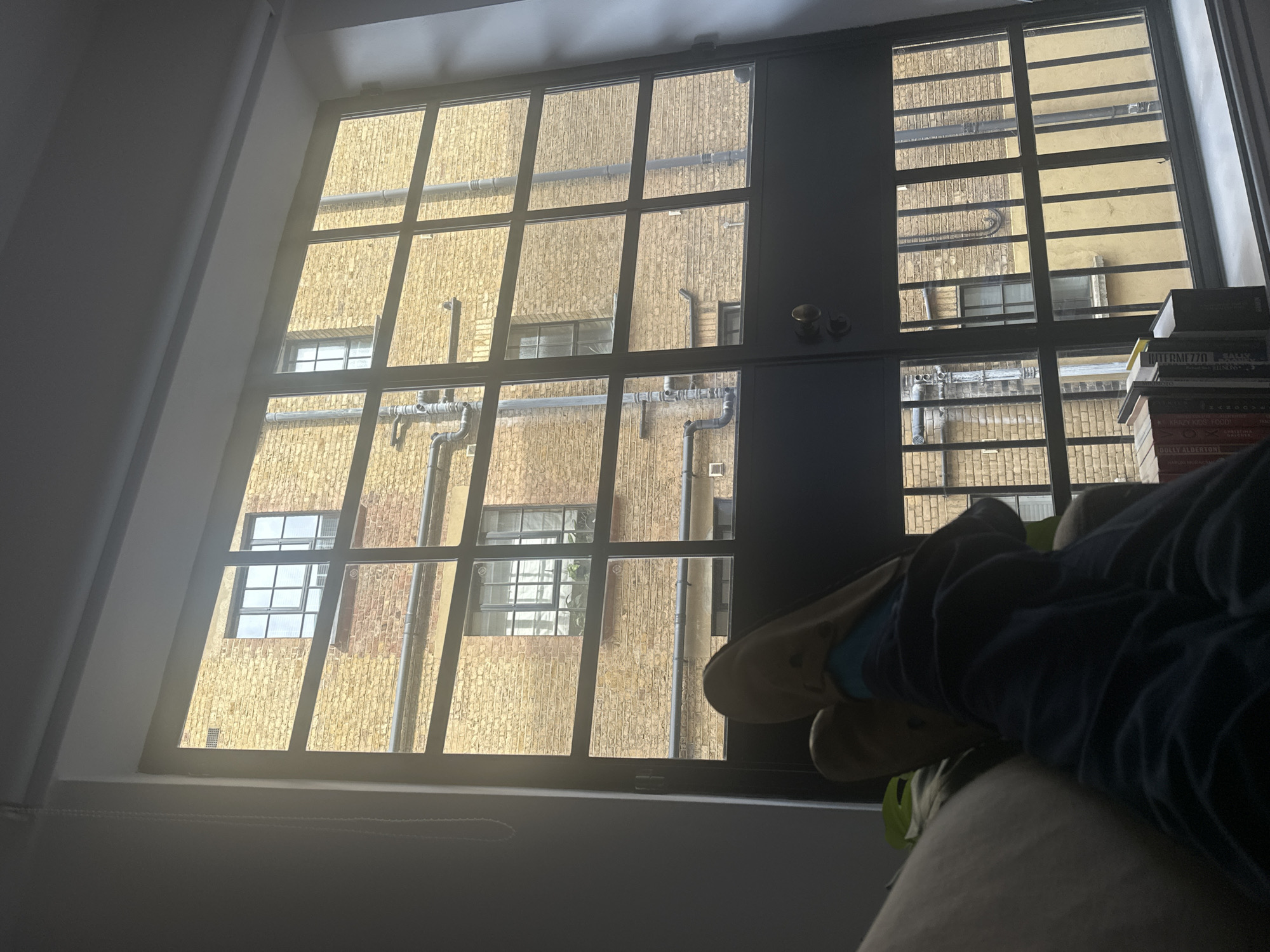The image shows a view from inside a room, looking out of a large, multi-paned window. Outside the window, a building with a brick facade is visible, featuring multiple windows and exterior pipes. The building appears to be in close proximity to the room's window. To the right of the window, a stack of books is partially visible. In the foreground, there is a person reclining, with legs visible wearing socks and slippers. The location may be in the city of London, United Kingdom, judging by the style of architecture of the building exterior.