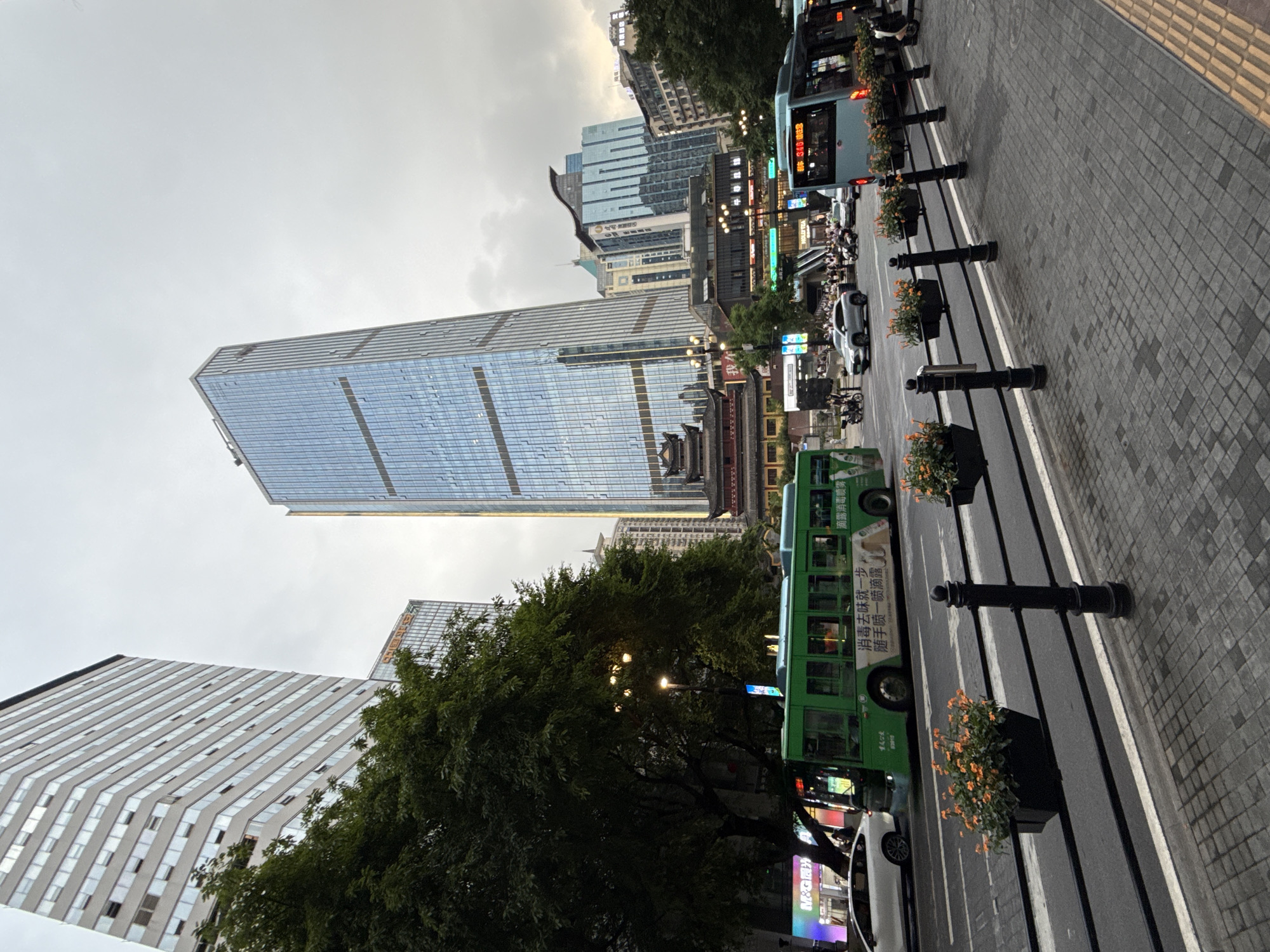 This is a street-level, slightly low-angle, wide shot of a busy urban intersection in Yuzhong District, China, likely during daytime given the overcast sky. The scene is dominated by modern skyscrapers and traditional Chinese architecture. In the foreground, a black metal fence with planters filled with orange flowers lines a pedestrian walkway paved with grey and tan bricks. The walkway is adjacent to a multi-lane road where traffic is flowing.
A bright green public transit bus is moving from left to right across the frame. It has Chinese text and an advertisement on its side. To the right of the green bus, a light blue bus is also moving in the same direction, with its destination displayed in red digital text. Several cars, including a silver SUV, are also visible on the road. Pedestrians are present on the sidewalk and crossing the street in the mid-ground.
Towering over the scene are several high-rise buildings. A very tall, modern skyscraper with a glass facade dominates the center-right of the image. To its left, a large, older, light-colored building with numerous windows rises prominently. In the mid-ground, a traditional Chinese building with a dark, tiered roof and red accents is nestled between modern structures. The sky is overcast and grey, suggesting it might be a cloudy day. The overall impression is of a vibrant, bustling city with a blend of contemporary and historical architectural styles.