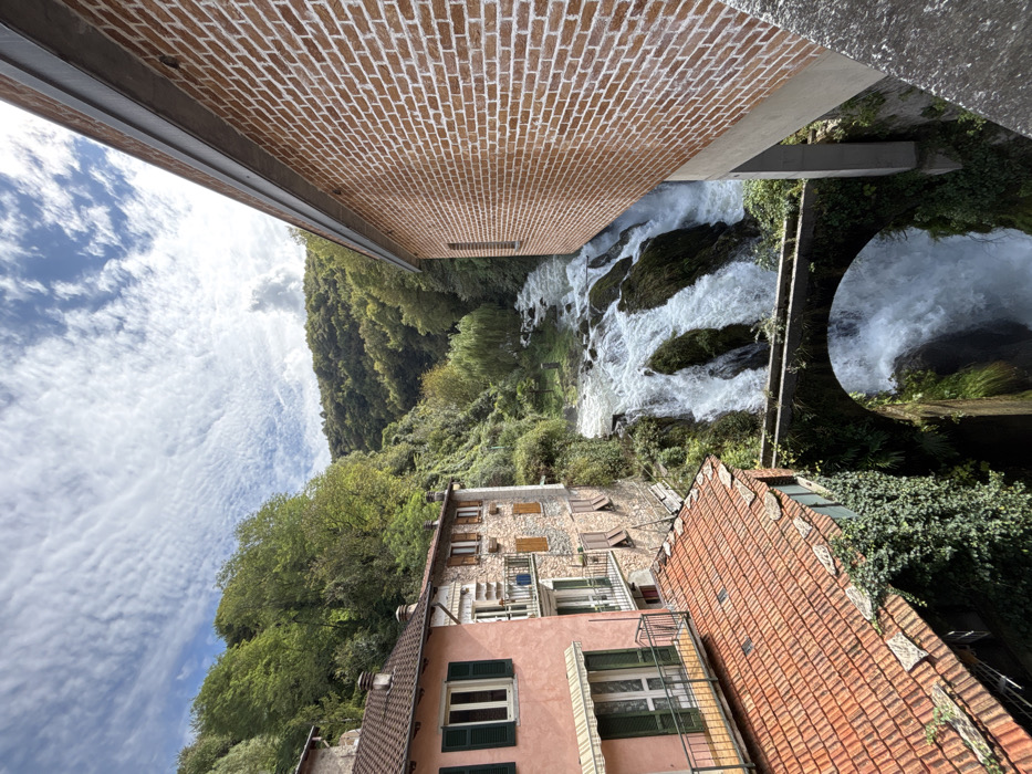 The image depicts a panoramic view of the Orrido di Nesso waterfall and its surrounding architecture in the city of Nesso, Italy. In the center, a powerful waterfall cascades down multiple tiers of dark rock formations, exhibiting foamy white water. An ancient stone arch bridge, partially covered in green ivy, spans the lower portion of the waterfall.
To the left, a multi-story building with a light peach-colored facade features green-shuttered windows and balconies with black metal railings. Its terracotta tiled roof is visible in the foreground. Adjacent to this, another multi-story structure with a stone facade, brown wooden-shuttered windows, and balconies is partially visible. Further up the slope, dense green trees and vegetation cover the steep hillsides, which form a valley around the waterfall.
On the right, a modern building with a reddish-brown brick facade and white mortar lines is prominently featured, extending from the mid-ground into the foreground. It has a small window and a grey roofline. The sky above is blue with scattered white, wispy clouds. No people or animals are visible in the scene. The perspective is from an elevated vantage point, looking down into the gorge.