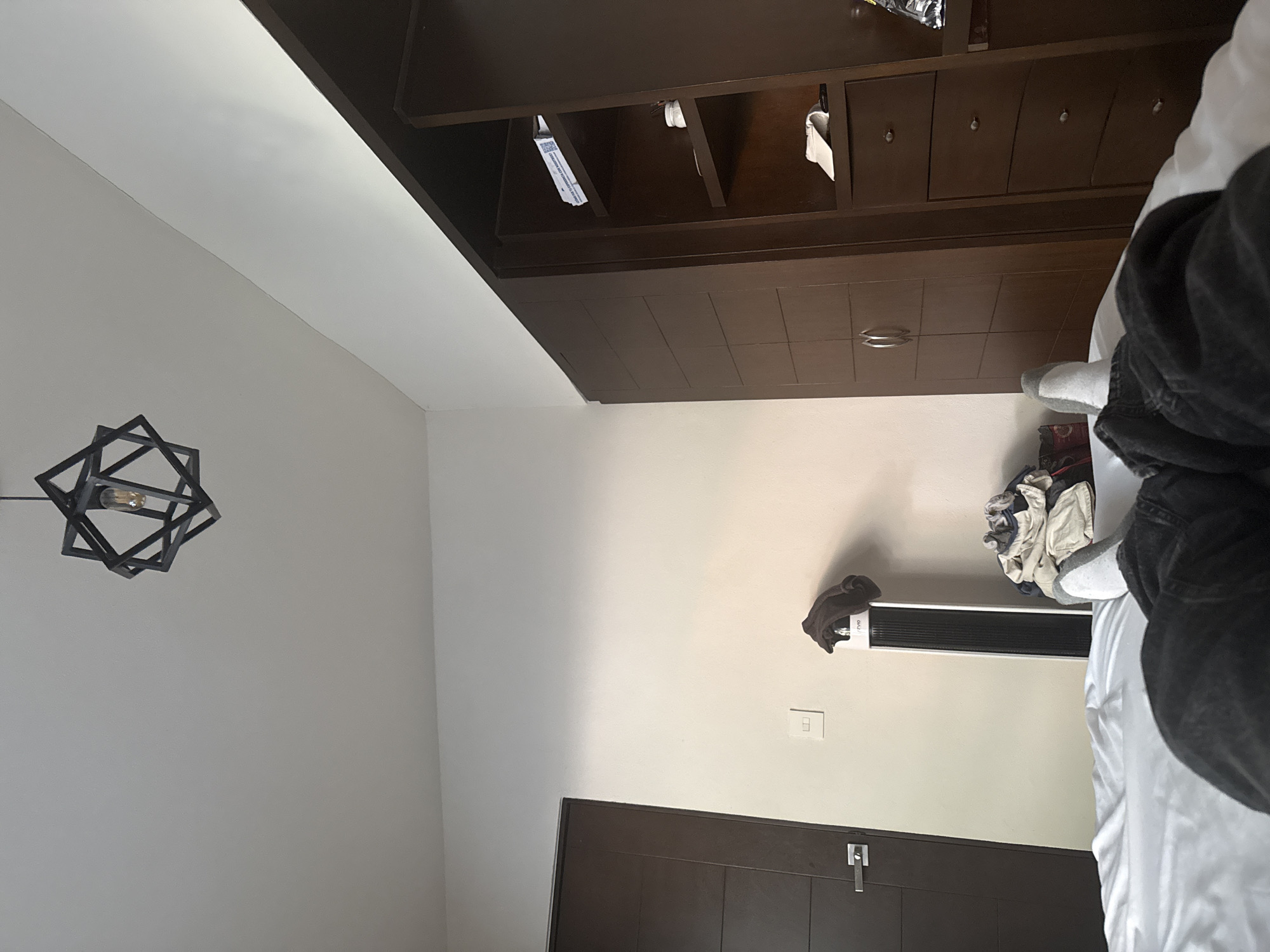 The image shows an indoor scene, potentially a bedroom. A dark brown wooden wardrobe is positioned along the top edge of the frame. Below the wardrobe, a section of a white wall is visible, featuring a geometric black light fixture on the left side. Further down, there is a wall-mounted white electric heater. A partially visible dark brown wooden door is at the bottom of the image. A person or people are located in the scene and appear to be sitting on a bed with a white comforter. Based on the style of the room and architectural elements, it could be inferred the location is in a residential area of Puebla, Mexico.