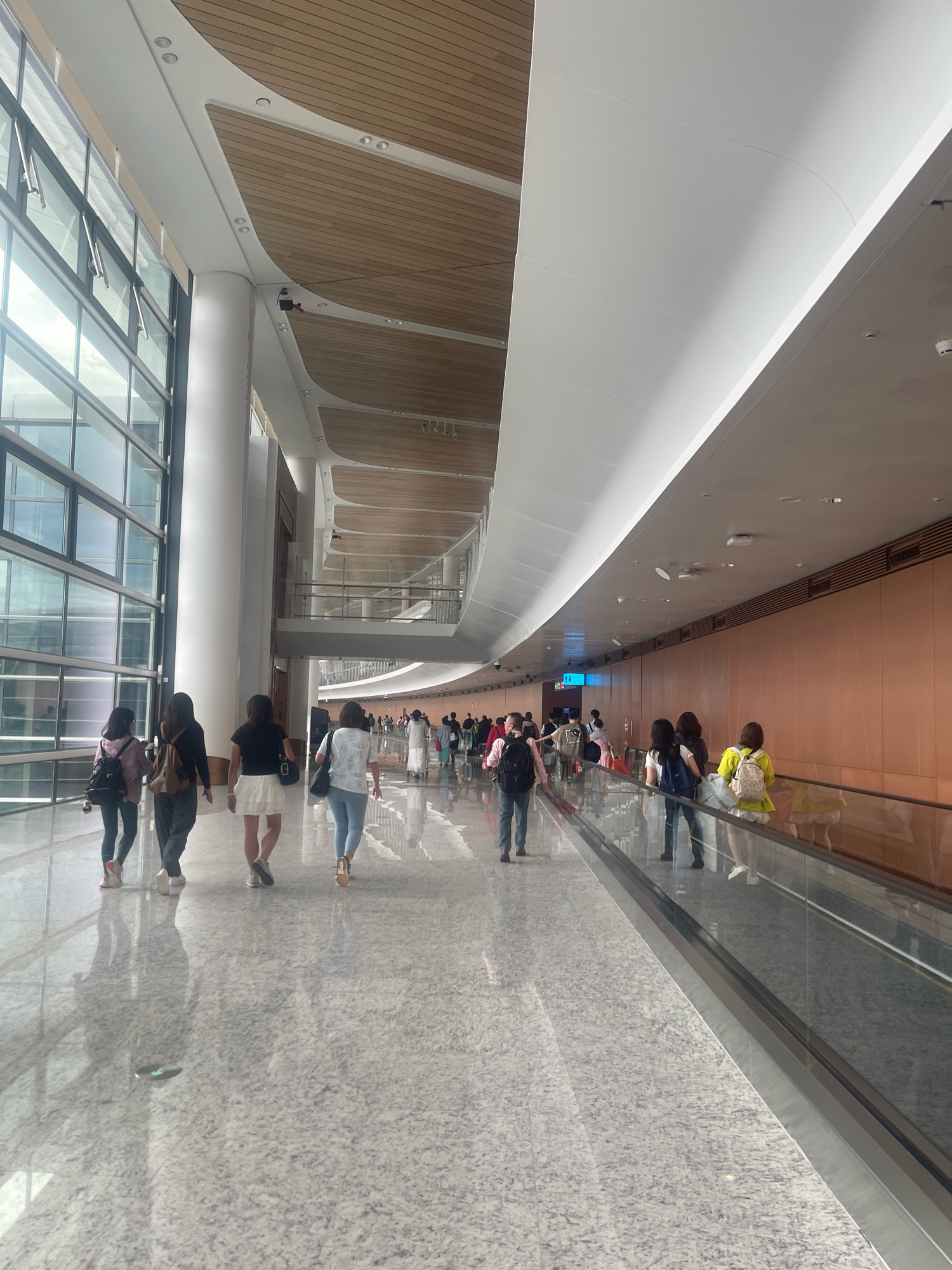 The image depicts the interior of a modern airport terminal in Baiyun District, China. A group of people are walking along a moving walkway, which is to the right of the frame. The walkway has a glass barrier separating it from the main concourse. The concourse floor is highly reflective, showing the reflections of the people and the architectural features above. To the left, a large expanse of glass windows allows natural light to enter, revealing a glimpse of the sky. The ceiling is a complex architectural design with curved white panels and wooden accents. Several people are walking in both directions on the concourse, some carrying luggage, suggesting travel. The overall environment is clean, spacious, and contemporary.