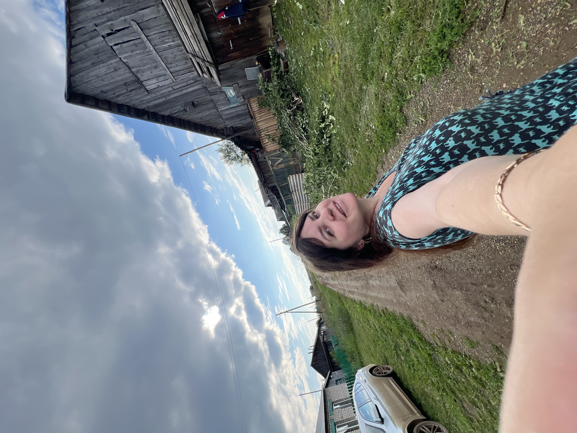 In Panteleevka, Russia, a woman takes a selfie on a dirt road. She is wearing a teal and black patterned dress and a gold bracelet. Behind her, a silver car is parked on the left, and a rustic wooden building stands on the right. Fallen branches and leaves are scattered on the ground in front of the building. The sky is partly cloudy, with patches of blue visible.
