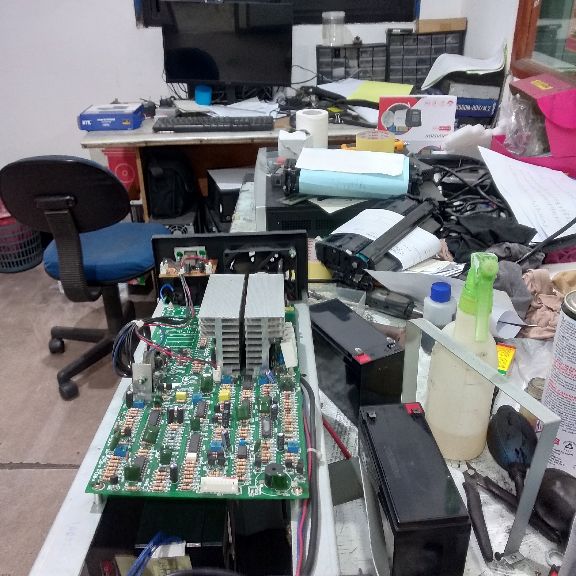 The image depicts a cluttered desk in what appears to be a workshop or office environment, likely located in Balaraja, Indonesia. The desk is covered with various electronic components, tools, and office supplies. In the foreground, a green circuit board with numerous resistors, capacitors, and integrated circuits is prominently displayed. Two large heatsinks are mounted on the board. Adjacent to the circuit board are two black batteries, one with red terminals.
Various other objects are scattered across the desk. To the left, a black office chair with a blue seat is partially visible. Further back, a computer monitor is visible, along with a keyboard and a blue cylindrical object. Several boxes and packaging materials are present, including a blue box labeled "NYK" and a white box with an image of security cameras. There is also a roll of white paper, a spray bottle with a green nozzle, and several tools, including a pair of pliers and a pair of wire cutters. Papers and documents are strewn across the surface. The overall impression is one of an active workspace where electronic equipment is being assembled, repaired, or tested. The lighting suggests it is daytime, and the environment appears to be indoors.