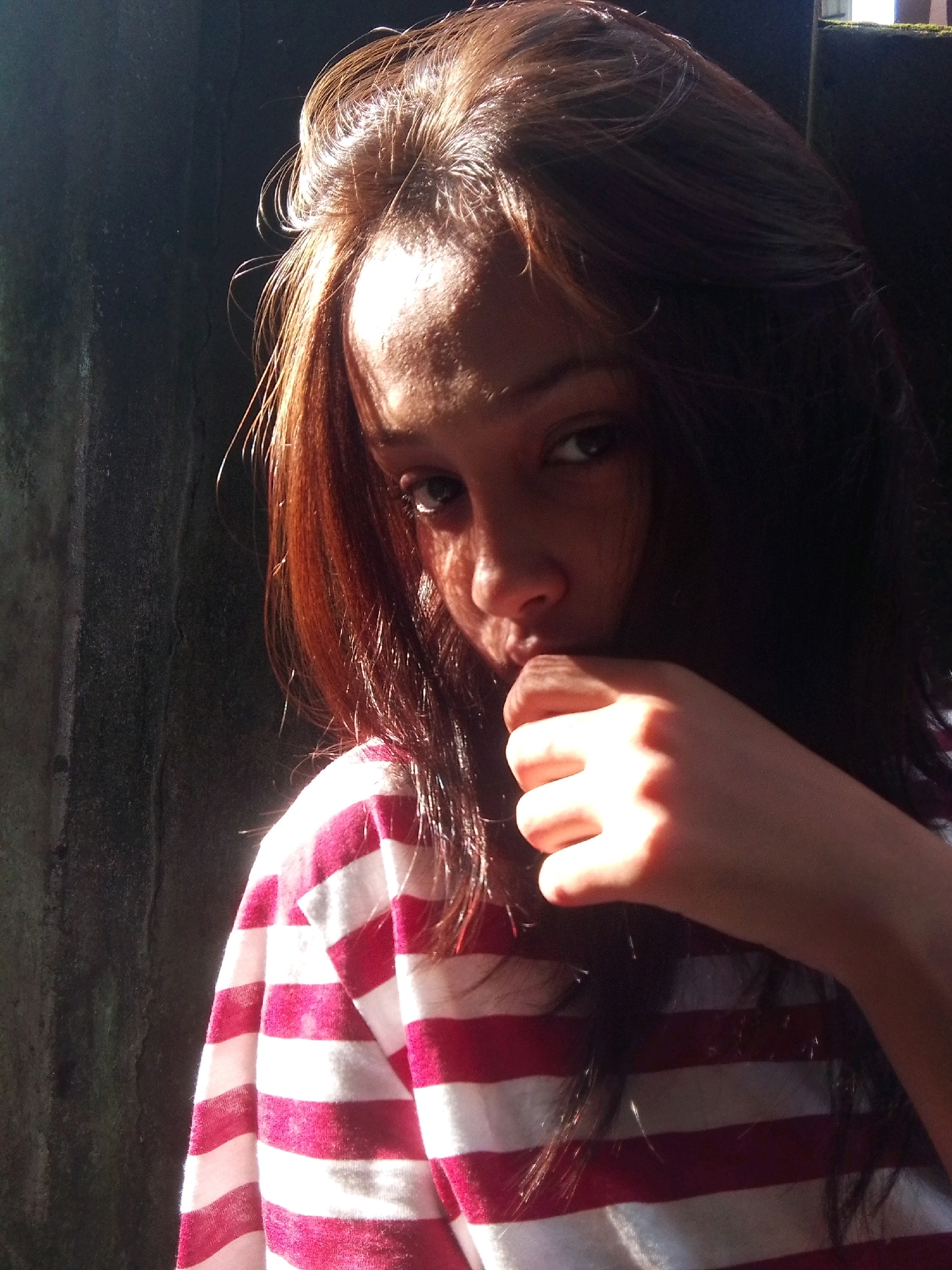 A young woman with long, dark hair is captured in a close-up portrait. She is wearing a red and white striped shirt. Her right hand is raised to her mouth, with her fingers touching her lips. Sunlight highlights her hair and face, creating a strong contrast with the dark background. The image appears to be taken outdoors, possibly against a textured wall. The woman is looking towards the viewer with a thoughtful expression.