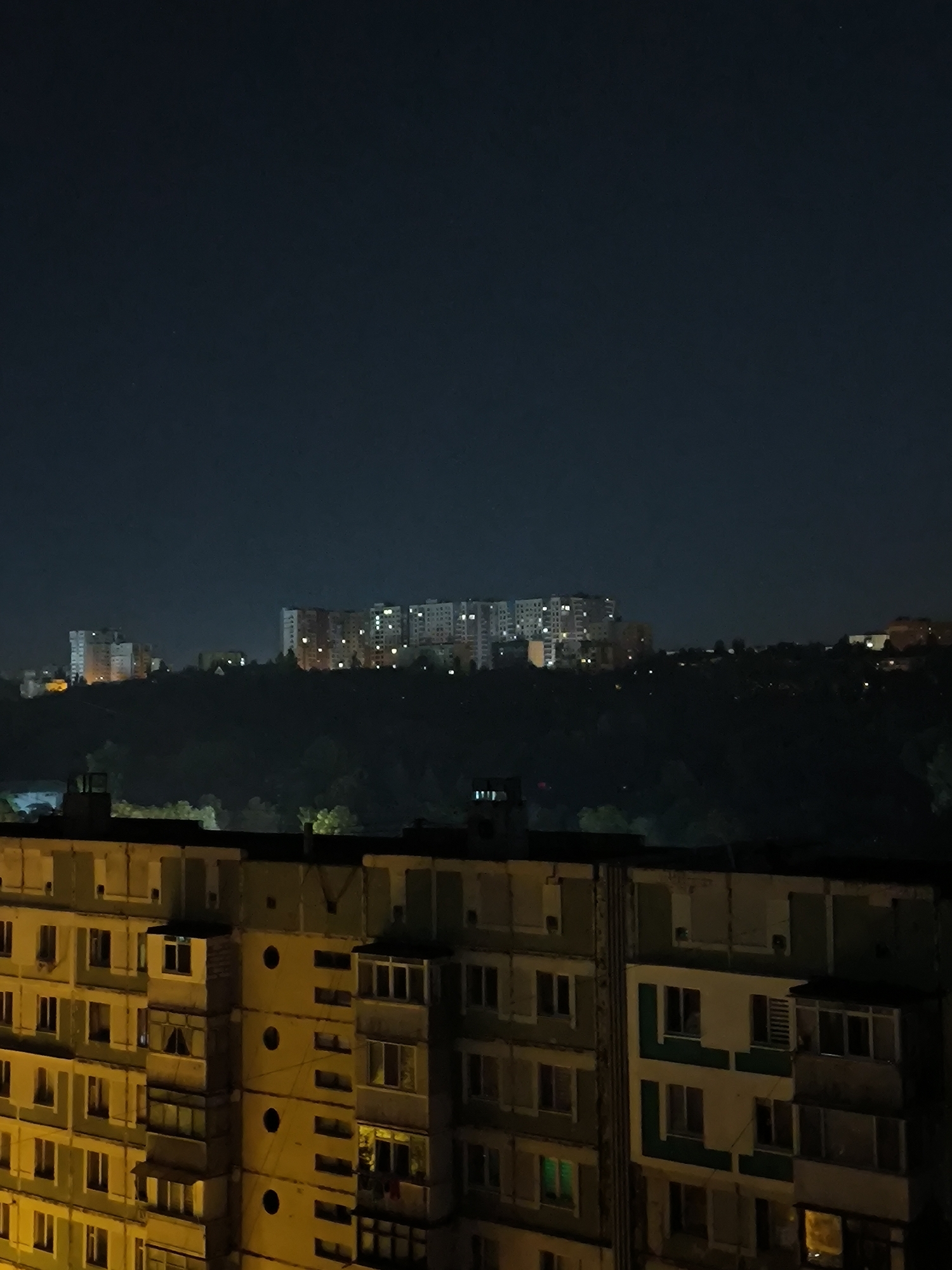 This is a nighttime, long-shot, exterior photograph of a residential area in Chișinău, Moldova. In the foreground, the image shows the facade of a multi-story apartment building, characterized by its weathered, light-colored exterior and numerous windows, some of which are illuminated. The building has a series of balconies and architectural details, including some protruding sections and circular windows.
In the mid-ground, a dark, tree-covered hillside is visible.
In the background, a row of taller apartment buildings is illuminated by interior lights, suggesting a densely populated urban area on a hill overlooking the foreground. The sky is dark blue, characteristic of a nighttime scene, with a few faint stars visible. The overall impression is of a typical urban night landscape in a large Eastern European city. The specific location within Chișinău is not identifiable from the visual cues alone.