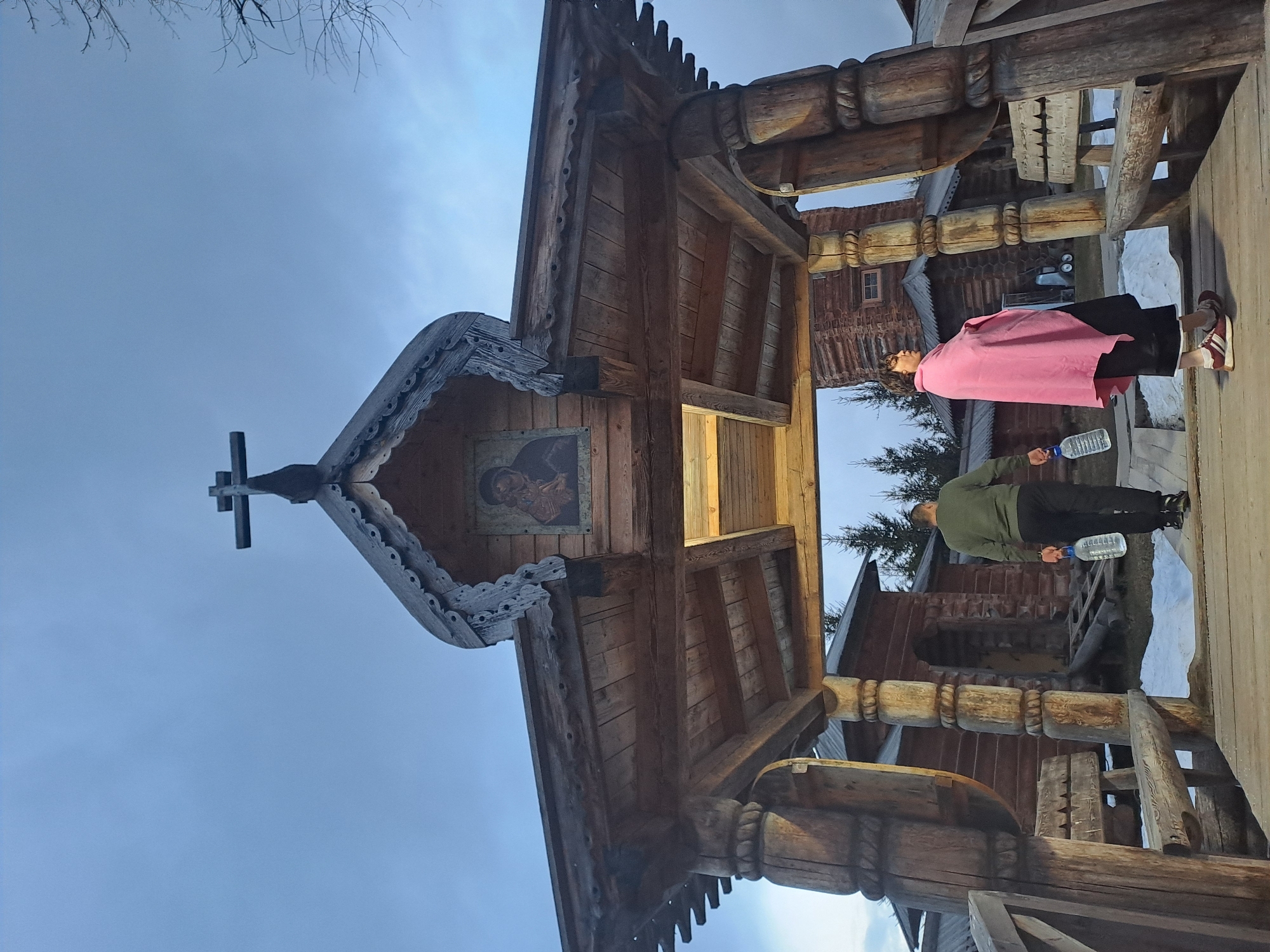 The image shows a wooden architectural structure, possibly an entrance or gate, with a cross at the top, adorned with an icon and intricate wood carvings. Two people are standing to the side of the structure: one wearing a pink shawl and black pants, the other in a green jacket and black pants. Both are holding water bottles. In the background, there are additional wooden structures suggesting a complex of buildings. This is located in the city of Vzglyadnevo, Russia.