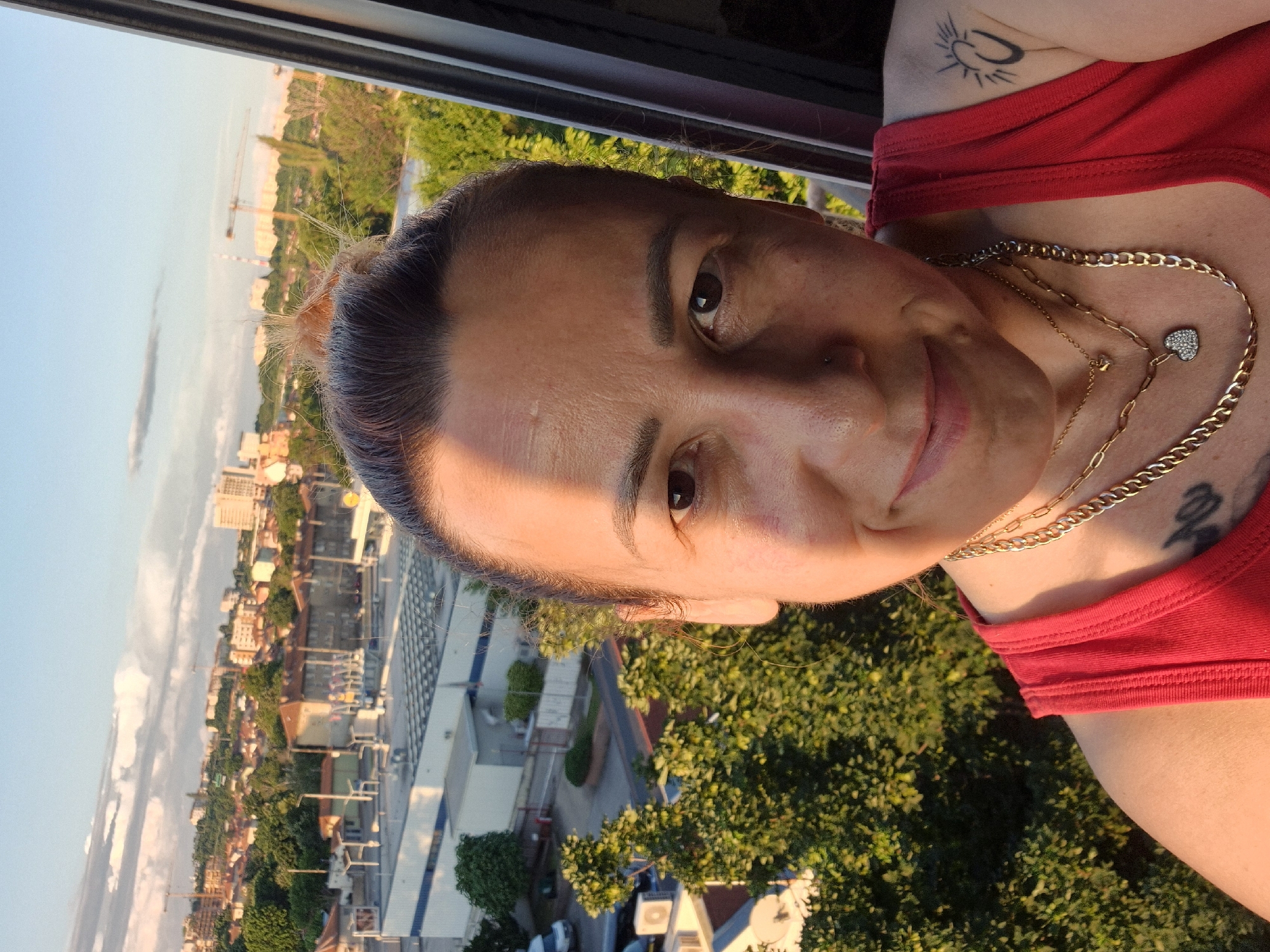 A close-up portrait features a woman seen from the chest up, with her head tilted significantly to the left, as if gazing out a window. She is wearing a red tank top and multiple gold-toned necklaces, one adorned with a small, heart-shaped pendant featuring tiny jewels. Tattoos are visible on her left shoulder, a stylized sun, and a darker, partially obscured design below her top. Her dark hair is pulled back, with fine strands framing her face. She has brown eyes, defined eyebrows, a subtle nose piercing on her right nostril, and a gentle smile.
Warm, golden sunlight illuminates the right side of her face and parts of her chest, indicating either sunrise or sunset. The background, seen through a dark window frame visible along the top of the rotated image, reveals an expansive urban or suburban landscape. Due to the image's rotation, the horizon appears vertical. The scene is filled with various buildings, lush green trees, and faint outlines of roads and vehicles. In the distant background, at least two large yellow construction cranes are visible against a light blue sky with a few wispy clouds. No discernible text is present.
