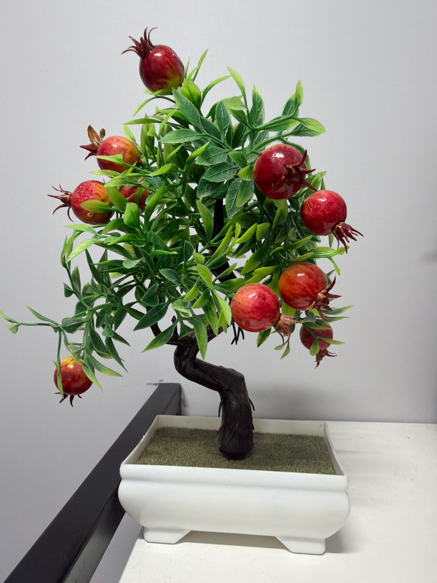 A small, artificial pomegranate tree with red fruits and green leaves stands in a white, rectangular pot filled with artificial grass. The pot has four short, square feet. The tree has a dark brown, twisted trunk and branches. The background is a plain, light gray wall. The pot rests on a white surface. A dark gray shelf or ledge is visible in the lower left corner. The image was taken indoors, likely in a room. There are no people or animals present. The image does not provide any information about its location in Vniissok, Russia.
