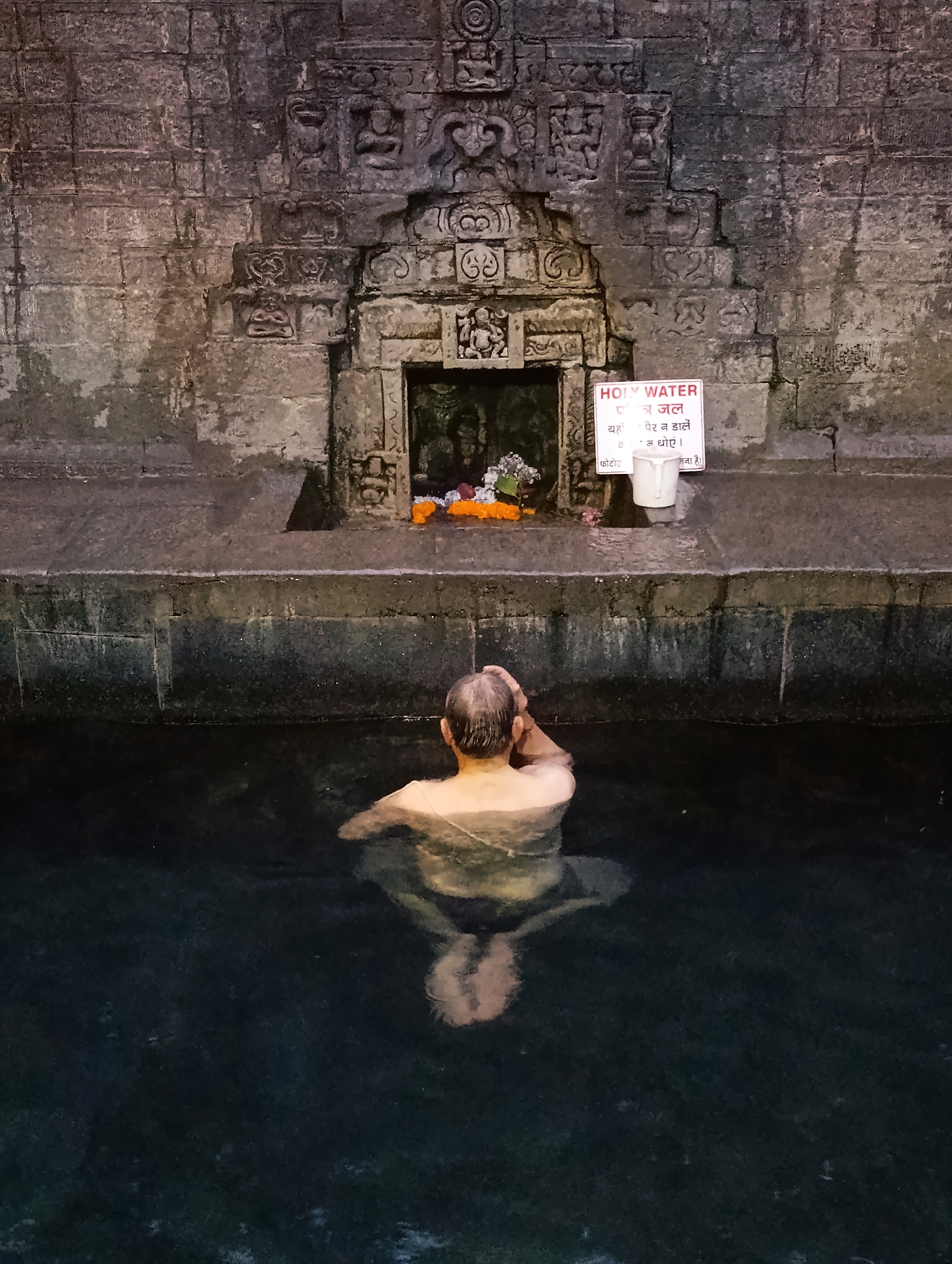 A man bathes in a dark, still pool of water at the Vashisht Hot Springs in Vashisht, India. The pool's edge is made of stone, and behind it is a carved stone wall with intricate reliefs. A small shrine with flowers sits in an alcove of the wall, and a white sign with Hindi text and the words "HOLY WATER" is posted to the right of the shrine. The man is facing away from the camera, with his head tilted back and his hands raised to his head. The water is calm, with ripples reflecting the carved stone structures above.