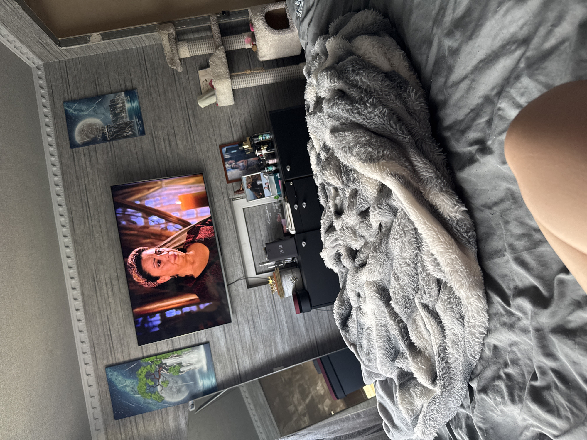 An indoor medium shot captures a cozy bedroom scene. The room is decorated with a gray, wood-plank style wallpaper. On the wall, a flat-screen television displays a woman with dark, curly hair and a patterned headband, looking downwards. To the left of the TV, a rectangular painting depicts a waterfall cascading from a tree with a large moon in the background under a starry sky. To the right of the TV, another similar-sized painting shows a lighthouse on a cliff overlooking the sea under a full moon and starry sky. Below the television, a black dresser holds various items, including framed photographs, decorative boxes, and bottles. A tall, floor-length mirror stands to the left of the dresser. In the foreground, a large, plush gray blanket is draped over a bed, with a person's leg visible at the bottom right corner of the frame. A multi-tiered cat tree with white fuzzy platforms is positioned in the background to the right. The lighting suggests it is daytime, with natural light filtering into the room.