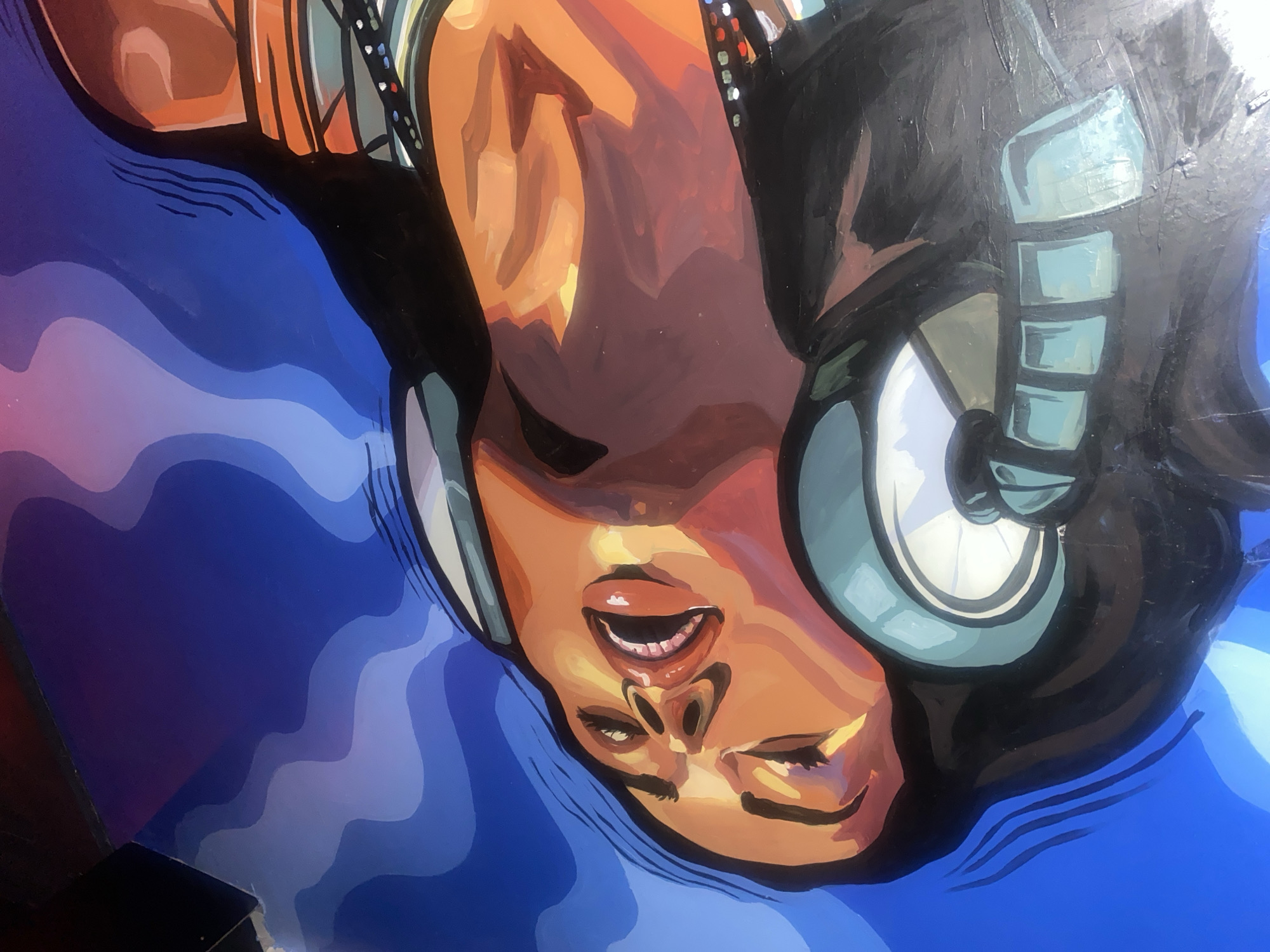 The image is a mural of a woman wearing headphones, with her eyes closed and mouth open as if singing or listening intently. The mural is rendered in a bold, colorful style with sharp lines and distinct color blocks. The woman's skin is depicted in warm tones of orange and brown, contrasting with the cool blue and purple background, which features flowing, abstract shapes that suggest sound waves or clouds. The headphones are a prominent feature, with teal and white accents. The overall impression is one of vibrant energy and immersion in music. This mural is located on a wall in Howayi, Nigeria.