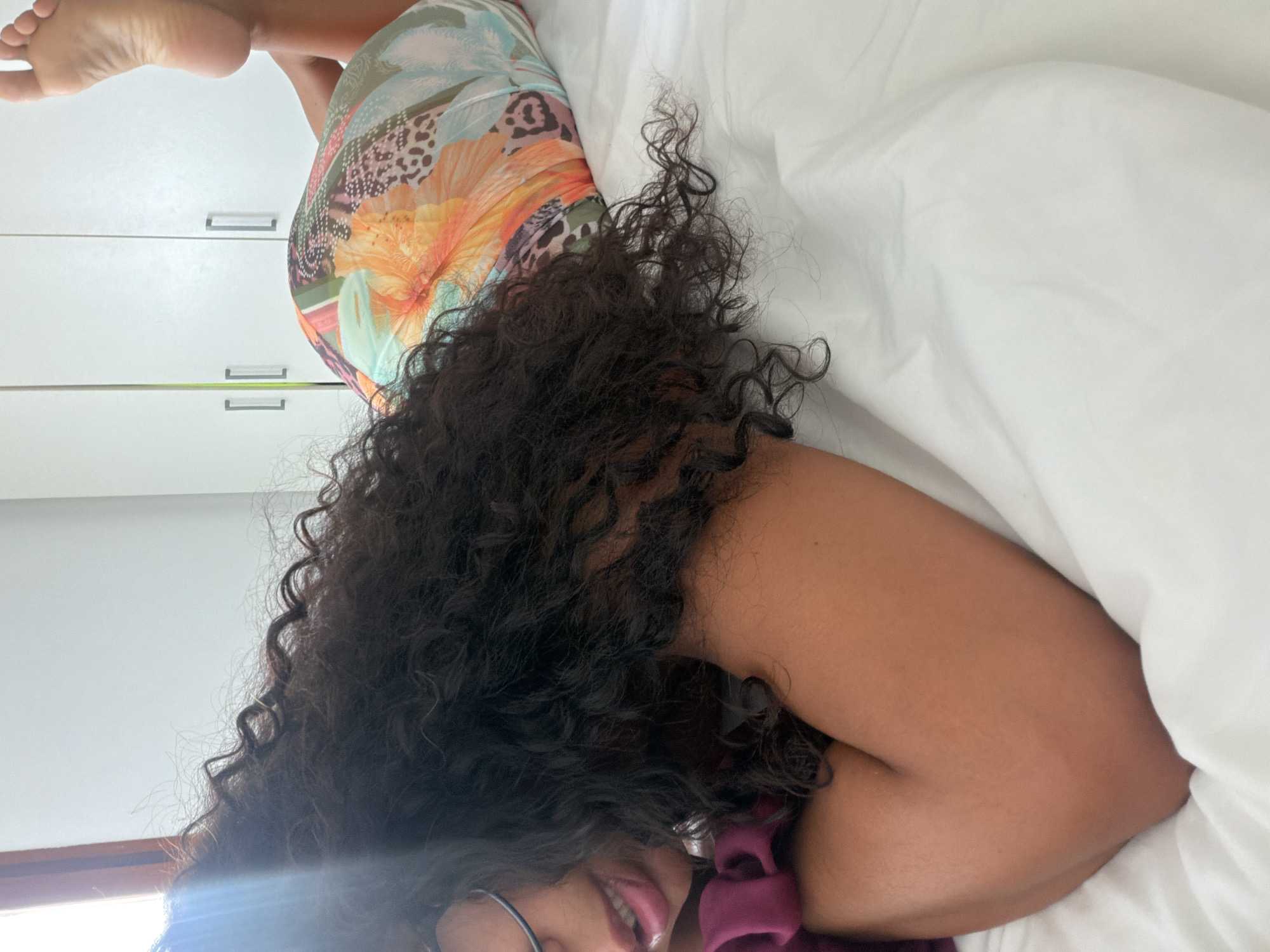 A woman lies on a white bed sheet in what appears to be a bedroom. She is wearing a floral patterned dress. The woman is positioned with her back facing upwards towards the viewer and her feet are elevated on a set of white cabinets. Her curly dark hair is spread across the pillow. The lower portion of her face is visible; she is wearing glasses and smiling. The location cannot be verified as Barcelona, Spain, with the current image.