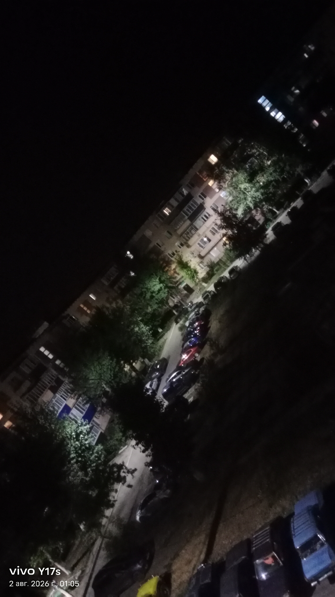 This is an aerial, tilted night-time photograph of a residential area in Rudny, Kazakhstan. The image captures a multi-story apartment building with lit windows, and a row of parked cars along a street. Trees and foliage are visible between the building and the street, illuminated by artificial light. The overall scene depicts a quiet urban environment at night.