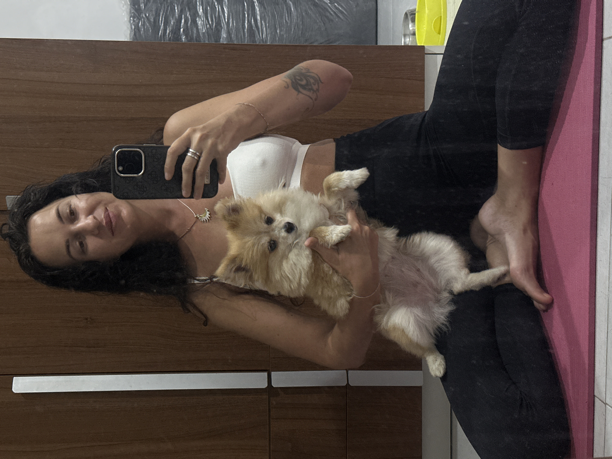 A woman with dark, wavy hair and a light complexion sits on a pink yoga mat, holding a small, fluffy Pomeranian dog in her arms. She is wearing a white crop top and black leggings. The woman is looking into a phone camera, held in her right hand, as if taking a selfie. She is wearing rings on her fingers and a necklace with a sun pendant. The background appears to be an indoor setting with wooden cabinets. There is a tattoo on her left arm. The Pomeranian is facing the camera with its mouth slightly open. The overall scene suggests a moment of casual relaxation at home. The location is the city of Kuta Selatan, Indonesia.