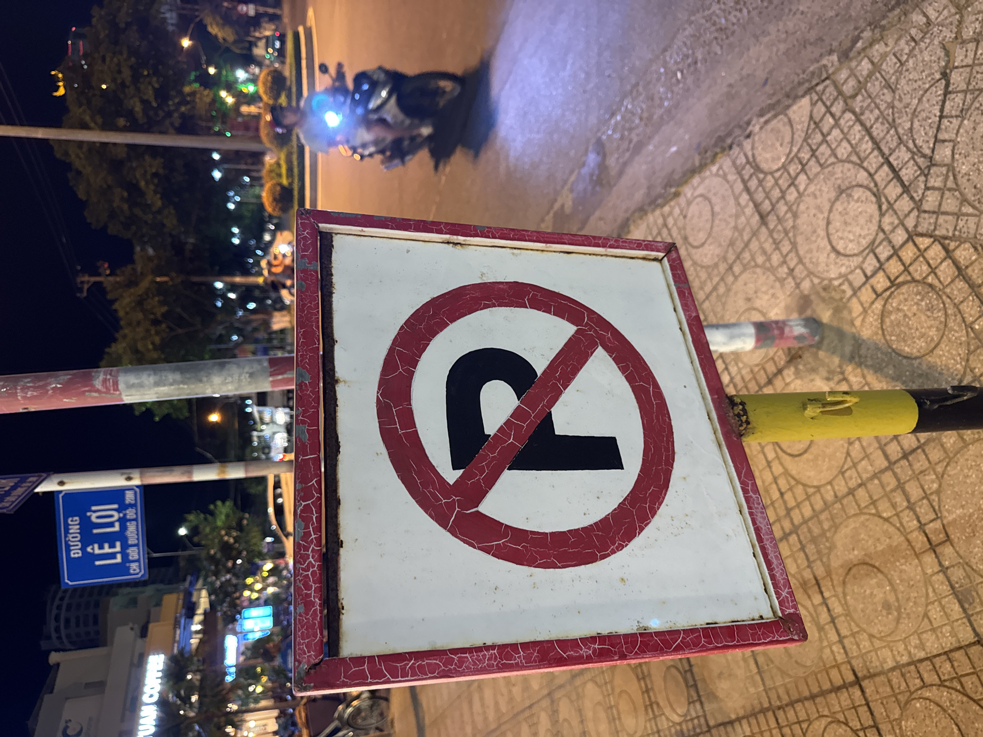 A close-up, eye-level medium shot captures a weathered "No Parking" sign on a street at night. The sign, a white square with a red circle and slash over a black "P," is mounted on a yellow and black pole. The red border of the sign is cracked and peeling, with visible rust. The pavement beneath the sign is made of square tiles with circular patterns. In the background, a street lamp illuminates a circular island in the road, where a person on a scooter with a bright headlight is passing by. A blue street sign in Vietnamese that reads "ĐƯỜNG LÊ LỢI" (Le Loi Street) is visible above and to the left of the "No Parking" sign. Buildings with illuminated signs and trees are also present in the background. The overall lighting suggests it is nighttime in a city.