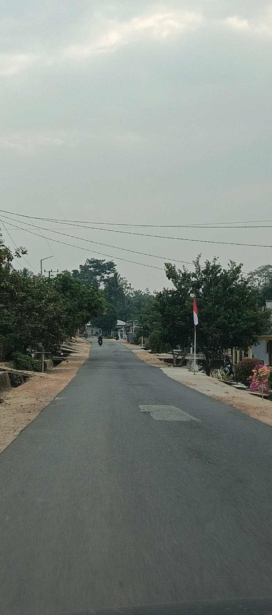 This is a vertical, eye-level shot of a paved road in Desa Puput, Indonesia, under a hazy, overcast sky. The road stretches into the distance, flanked by vegetation and buildings. On the left side, a sandy embankment with drainage channels lines the road, bordered by lush green trees. On the right side, trees and some residential structures are visible, including a house with a red and white flag flying from a pole. A few motorcycles are seen on the road, one in motion in the middle distance, and at least one parked on the right side. Power lines crisscross the sky above the trees. The overall atmosphere is subdued due to the cloudy weather.