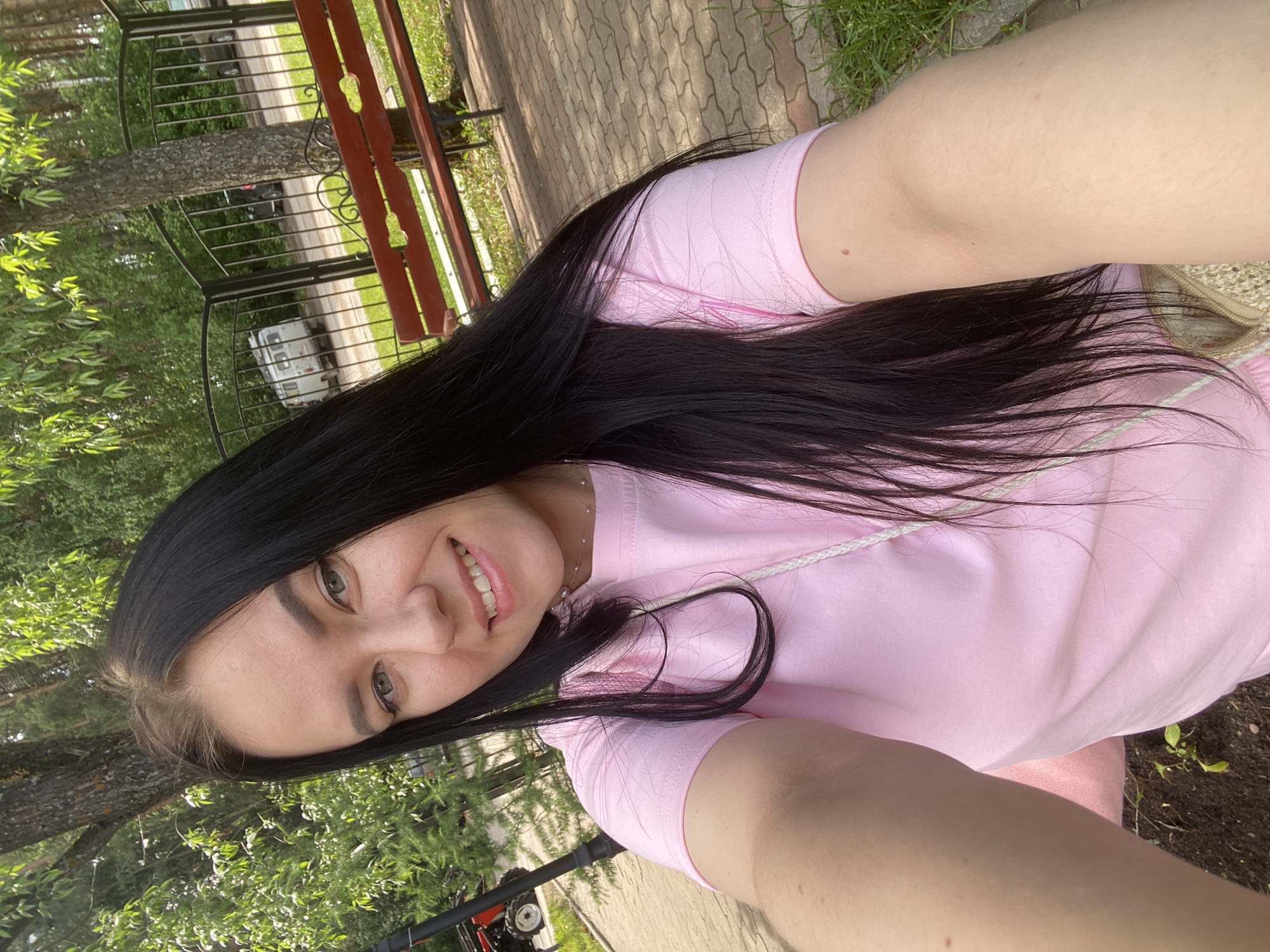 A selfie photograph captures a smiling young woman outdoors in Chaikovsky, Russia. She has long, dark hair that flows over her shoulders and part of her chest, and she is wearing a light pink t-shirt with a white drawstring detail at the neckline. A delicate, light-colored necklace with a small pearl or bead is visible around her neck. Her arm and shoulder are prominent in the foreground, indicating the selfie perspective.
The background reveals a lush green environment, primarily composed of trees and dense foliage, suggesting a park, garden, or well-vegetated area. A dark, ornate metal fence or gate with a red wooden top rail separates the foreground from a paved path or road. Beyond the fence, a white camper van is partially visible, parked on or near the road. The ground in the immediate foreground consists of grey and reddish pavers interspersed with patches of grass and dirt. A small piece of red and black machinery, possibly a lawnmower, is faintly seen in the lower left corner.
The scene is brightly lit, indicating a clear, sunny daytime. The woman's cheerful smile conveys a positive and relaxed mood. No visible text is present in the image.