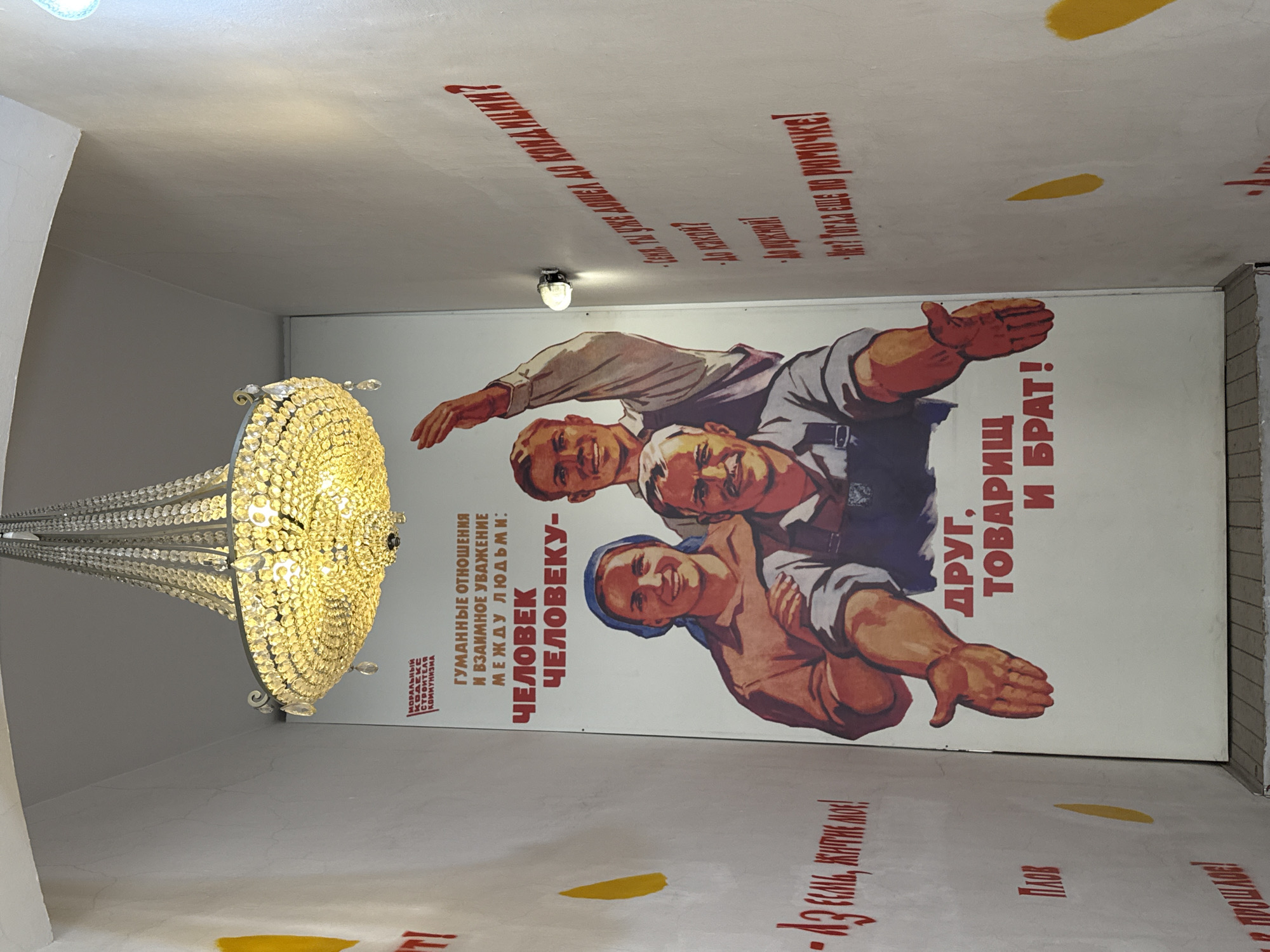 The image shows a section of a building interior in Moscow, Russia, featuring a Soviet-era propaganda poster on the wall. The poster depicts three smiling workers of different ethnicities embracing. The text on the poster promotes "humane relations and mutual respect between people" and states "Man to Man - Friend, Comrade, and Brother!". Red text is stenciled on the ceiling above the poster. A large, ornate chandelier hangs to the left of the poster. There are yellow paint marks on the ceiling.