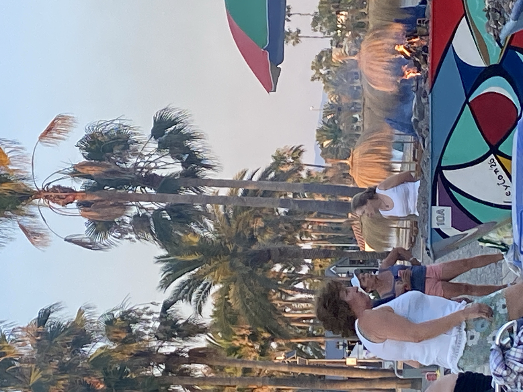 In Marbella, Spain, a group of people are gathered near a beach bonfire. A woman with curly brown hair, wearing a white tank top and a floral patterned skirt, stands in the foreground. Behind her, two other women are visible. One, wearing a blue tank top and pink shorts, holds a cup. The other woman, in a white tank top, is looking towards the bonfire. The bonfire is situated on a colorful, decorated structure with the number "10A" visible. In the background, palm trees line the beach, and a brightly colored umbrella stands to the right. The scene appears to be set during a sunset, with a warm, soft light illuminating the area.