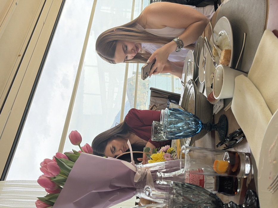 The scene depicts two adult females seated at a dining table inside a restaurant with large windows in Budva, Montenegro, providing a view of the Adriatic Sea and a rocky landmass, consistent with Sveti Nikola Island, under an overcast sky. The woman on the left, with long dark hair, wears a dark red top and is looking down at an object in her hands. The woman on the right, with long light brown hair, wears a white sleeveless top and is actively engaged with a silver-colored smartphone held in both hands; a watch is visible on her left wrist. The wooden table is set with gray placemats, multiple partially cleared plates with food remnants, cutlery, various drinking glasses including two blue textured goblets, a clear bottle of dark carbonated beverage, and a tall clear glass containing a dark liquid with an orange slice. A large bouquet of pink tulips wrapped in light purple paper and tied with a white ribbon, along with a smaller arrangement of yellow and pink flowers, serve as centerpieces.