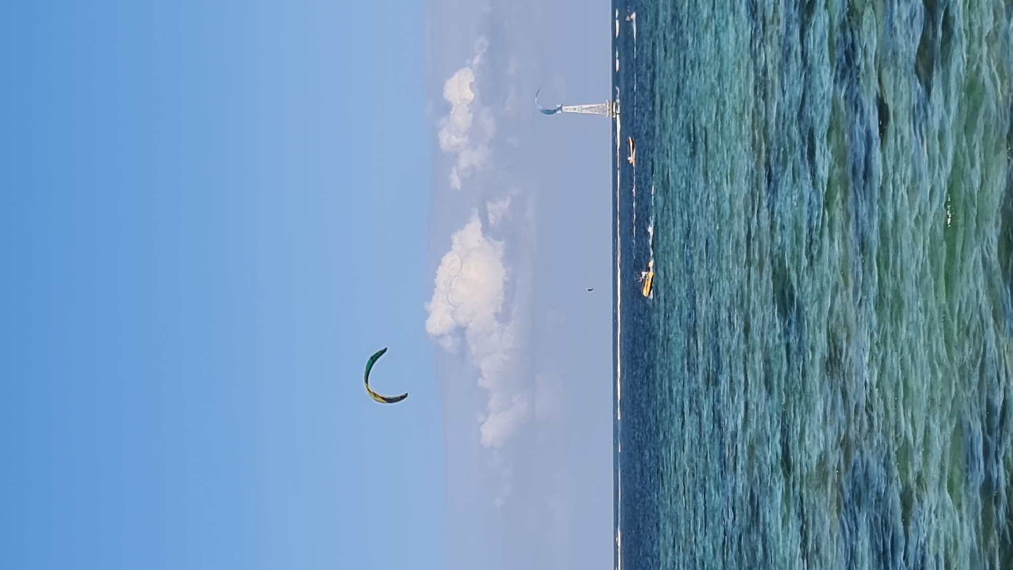 The image depicts a bright, sunny day at a coastal location, likely in or near Denpasar, Indonesia. The scene is dominated by the ocean and the sky. The water in the foreground is a vibrant turquoise, with visible waves and ripples. Further out, the ocean transitions to a deeper blue. A thin white line of breaking waves suggests a reef or shallow area in the distance.
Against the clear blue sky, several elements are visible. A large, crescent-shaped kite, colored with green, yellow, and blue, is suspended high in the air, indicating kiteboarding or a similar activity. In the mid-ground, a small yellow boat with a white cabin is moving across the water, leaving a white wake. Further to the right, a tall, slender structure, possibly a wind turbine or a radio tower, stands on the water. There are other, less distinct boats and kites scattered across the horizon. Fluffy white clouds are present in the sky, adding texture and depth. The overall impression is one of a recreational or sporting activity taking place on a beautiful day by the sea.