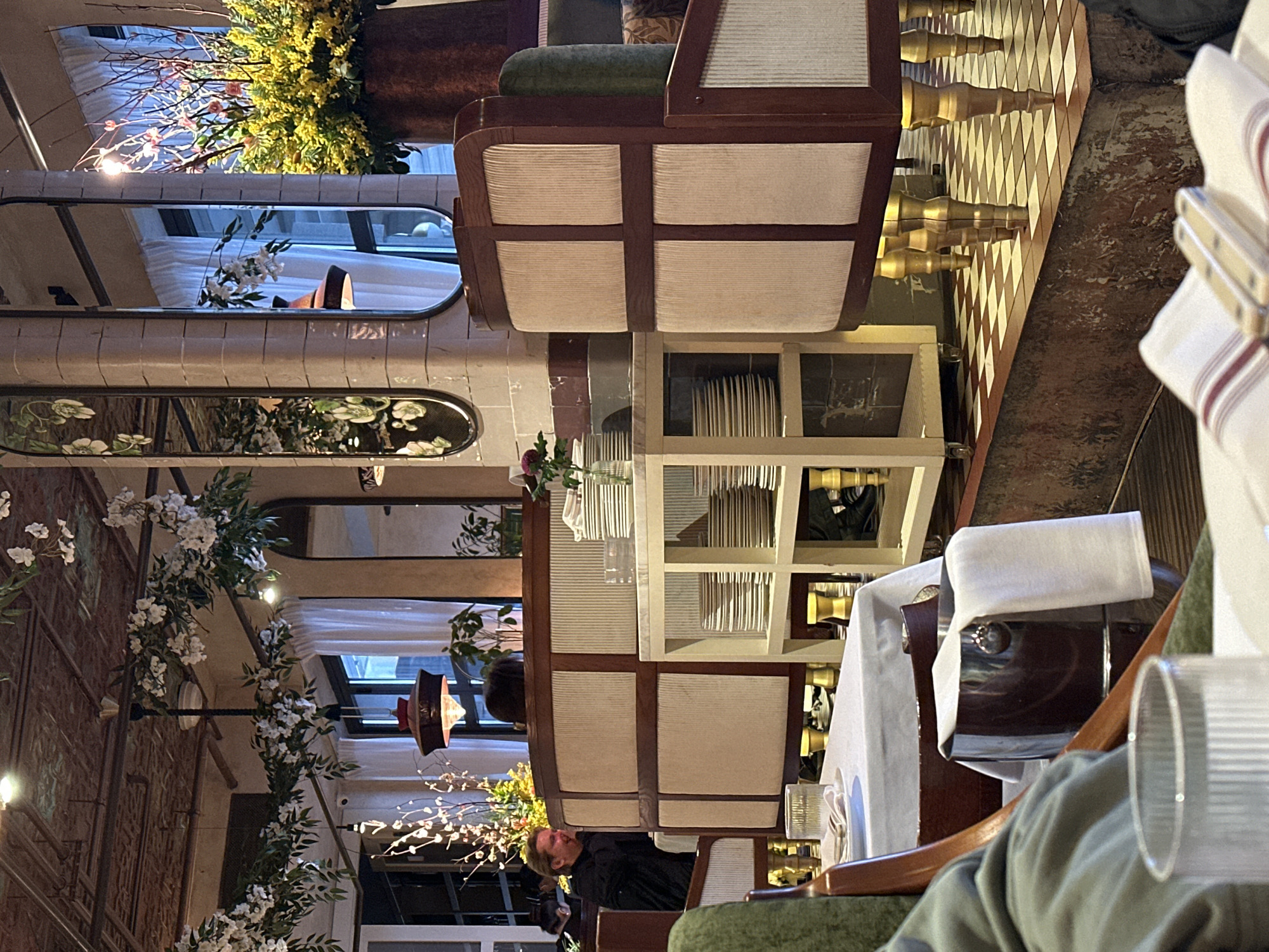 The image captures the interior of a restaurant in Moscow, Russia. It features a dining area decorated with floral arrangements, including white and yellow flowers. The restaurant has a unique interior design with mirrors, patterned flooring, and tiled walls. Furniture includes upholstered chairs and tables set for dining. A person is seated at one of the tables. Tableware, such as plates, glasses, and napkins, are visible on the tables.