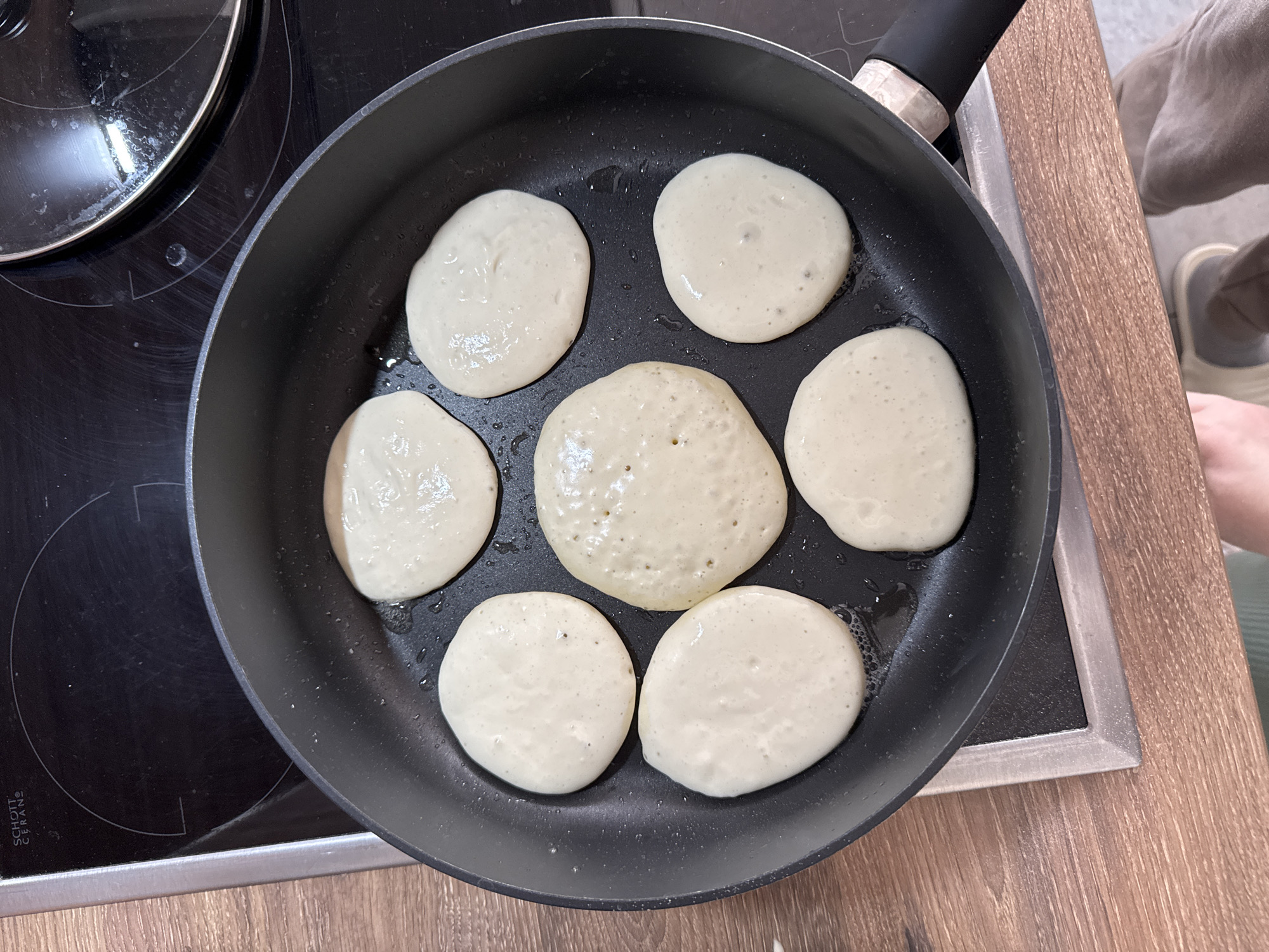 In an overhead shot, an electric stovetop burner is visible, with a black frying pan on top. Seven pancakes in batter form are in the pan, arranged in a circular pattern with one pancake in the center. The pancakes are pale yellow and have a smooth, slightly bubbly surface. The stovetop is black and glossy, with circular markings and text that appears to read "SCHOTT CERAN". To the left of the stovetop, a wooden countertop is visible, with a person's legs and feet wearing casual clothing and shoes in the bottom left corner. The scene appears to be indoors, likely in a kitchen.