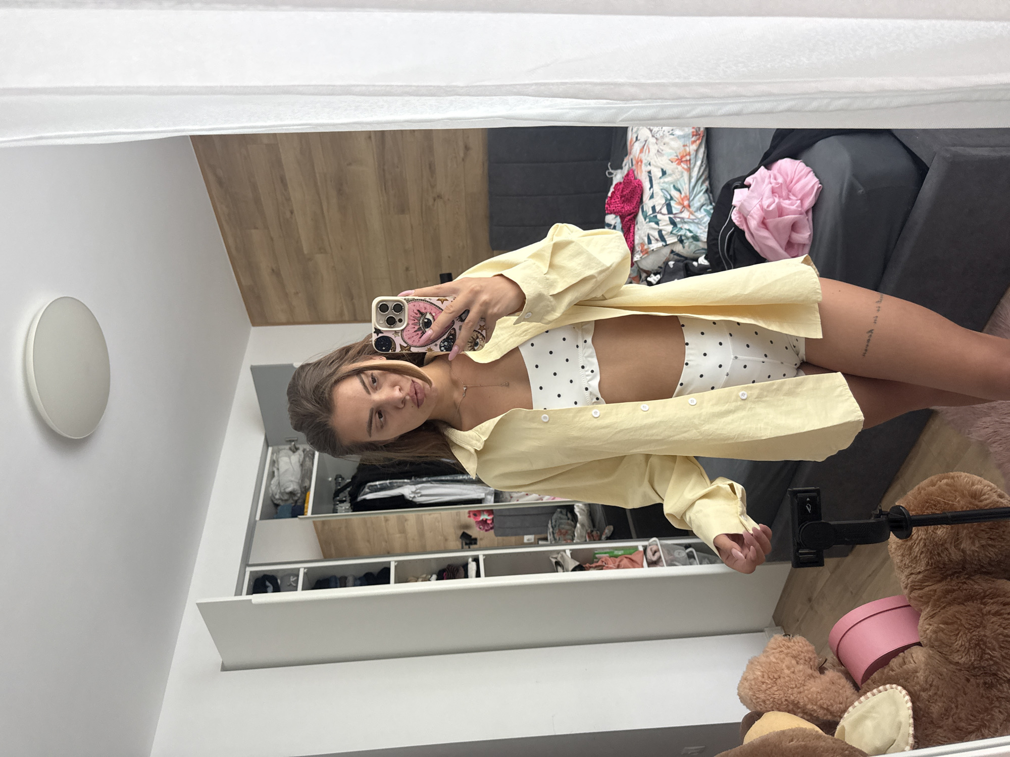 A young woman is taking a mirror selfie in a bedroom. She is wearing a yellow button-down shirt over a white polka dot bikini top and bottoms. Her hair is brown and pulled back. She is holding a phone with a heart-shaped case in her right hand. The room appears to be a bedroom with a large mirror, a closet filled with clothes, a bed with floral patterned bedding and pink clothes on it, and a large stuffed teddy bear. The room has a light-colored ceiling and walls, with a wooden accent wall behind the bed. There is a white curtain on the right side of the image. The photograph appears to have been taken indoors. The location is not specified in Plovdiv, Bulgaria.
