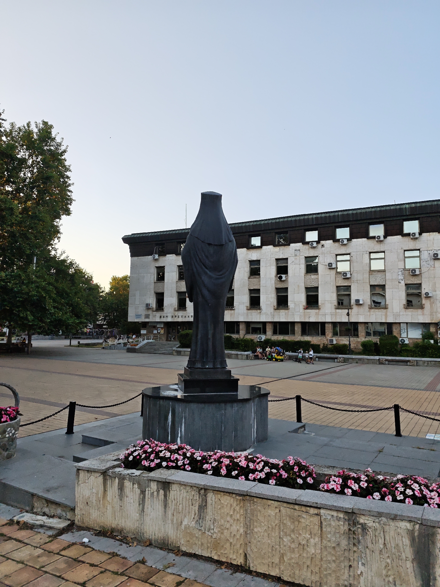 The image shows a public square in Lovech, Bulgaria. In the foreground, there is a dark, abstract statue, likely made of stone or metal, with a draped form. The statue stands on a circular base, which appears to be part of a fountain, though no water is visible. In front of the statue, there are two stone planters filled with bright pink flowers. The square itself is paved with light-colored and darker rectangular stones.
Behind the statue, a multi-story building dominates the background. It has a light-colored stone facade, with numerous windows and air conditioning units visible. The building's architecture suggests it is likely a public or administrative building. There is a sign above the entrance with Cyrillic text, which reads "ДОМ ПРЕДСЕДАТЕЛ" (Dom Predsedatel), meaning "House of the Chairman" or "Chairman's Building."
Several people are scattered throughout the square. In the middle ground, a small group of people are seated on benches and a low wall in front of the building. Some children are visible playing with a yellow toy vehicle. Further in the background, more people are seen sitting on benches and walking around the square.
To the left of the frame, a large tree with green foliage is present, suggesting a park-like setting. The sky is clear and pale, indicating it is likely late afternoon or early evening. The overall scene depicts a tranquil public space in a Bulgarian city.