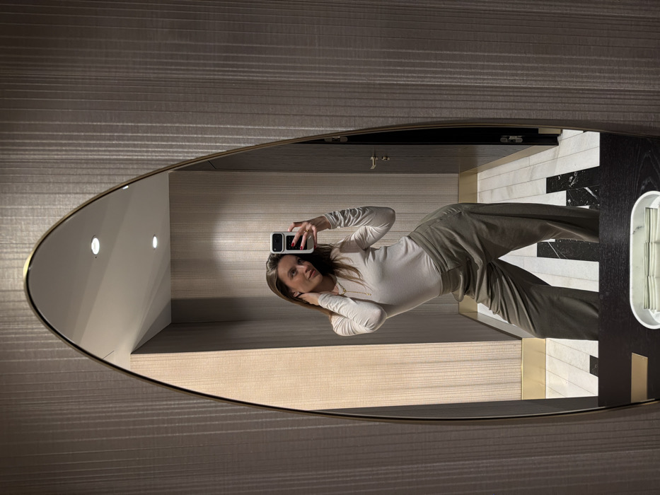 A female subject is depicted taking a mirror selfie in an indoor setting located in Budva, Montenegro. The subject, with long brown hair, wears a light-colored long-sleeved top, olive green wide-leg trousers, a gold-colored necklace, and small stud earrings. Her left hand holds a white-cased smartphone to capture the image; red nail polish is visible on her fingers. Her right hand is positioned near the back of her neck. The subject is reflected in an arched, gold-framed mirror. The reflected background consists of walls covered in textured, vertically striped beige wallpaper. The ceiling is white with two recessed circular light fixtures. The floor exhibits a distinctive geometric pattern of alternating black and white rectangular tiles. A dark-colored door with a metallic handle is visible in the upper right portion of the mirror's reflection. A dark counter is visible in the lower foreground, supporting a white oval container, likely a tissue dispenser.