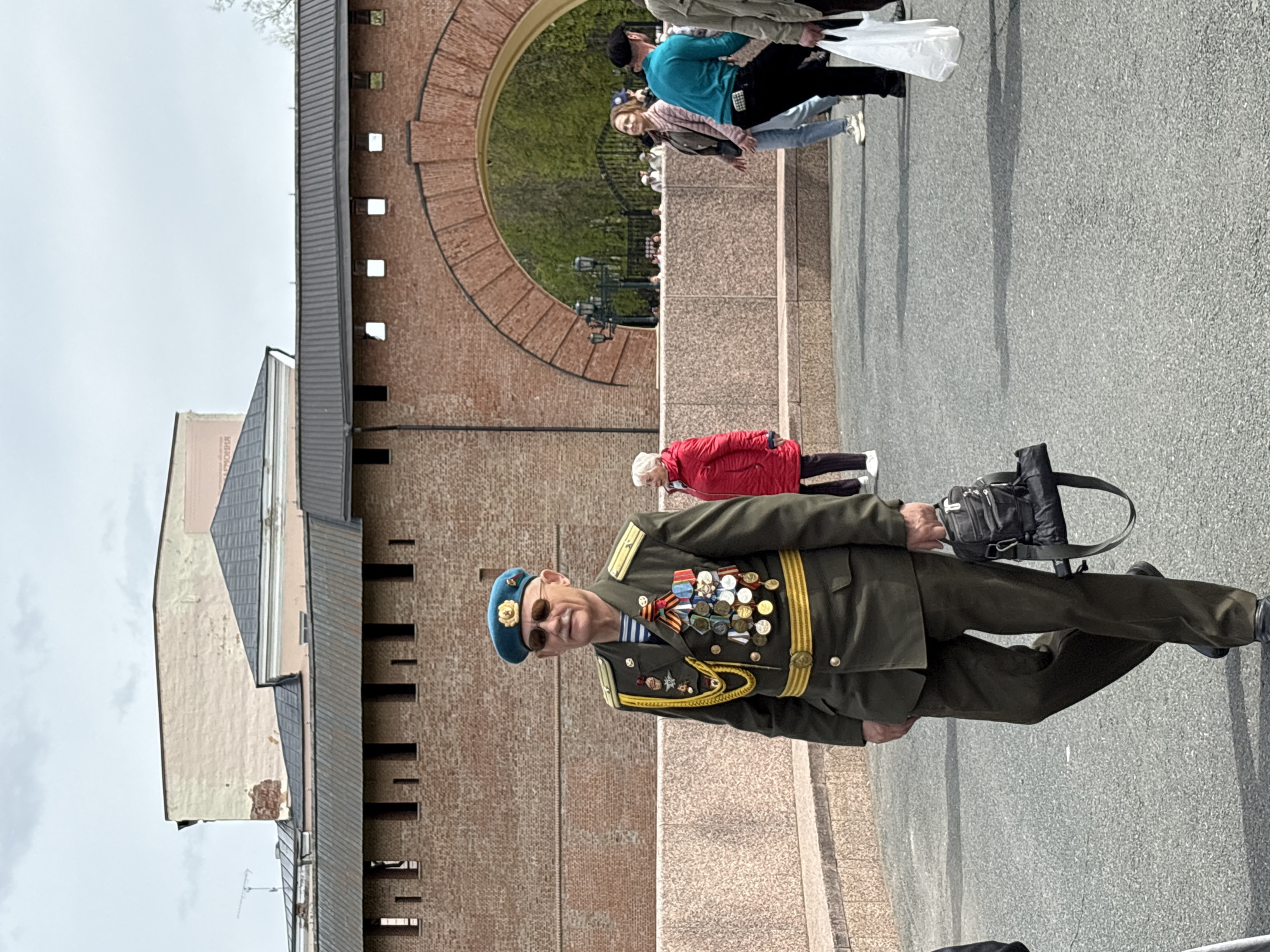 The image shows a man in a military uniform walking on a street near a brick wall with an arched opening. The man is wearing a green military uniform adorned with numerous medals and a blue beret. He is holding a black bag. In the background, there are other people walking, and architectural details suggest an urban environment. This image was taken in Veliky Novgorod, Russia.