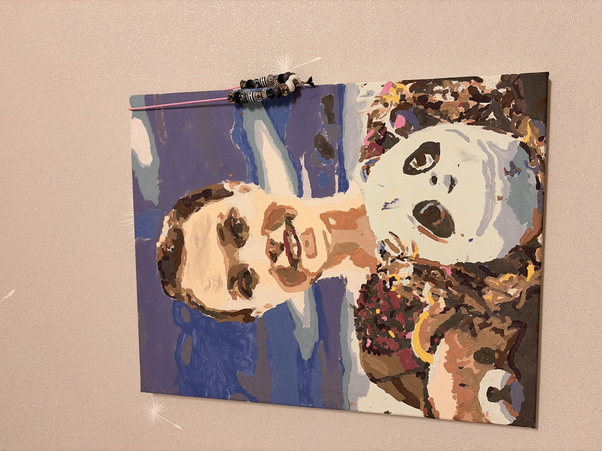 A painting of a person holding a panda is displayed on a wall. The painting is in a cubist style with bold blocks of color. The person in the painting has short brown hair and is smiling. They are wearing a brown shirt. The panda is white with black markings. The background of the painting is a blue sky with white clouds. The painting is hanging on a light pink textured wall. A pink string with beads is attached to the top right corner of the painting and hangs down the side. There are two white dandelion-like designs on the wall to the left of the painting. The painting appears to be in a domestic setting. There is no identifiable information about Leninogorsk, Russia in the image.