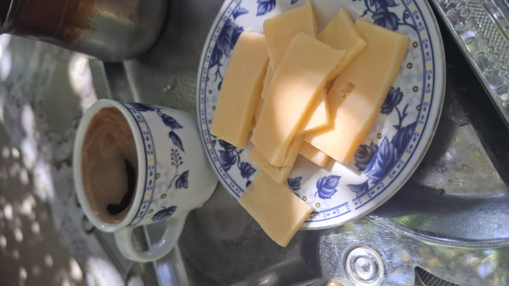 The image shows a close-up of a plate of sliced cheese and a cup of coffee, likely in Шұбарсу, Kazakhstan. The plate has a blue and white floral pattern. The coffee is dark and steaming. In the background, there's a glimpse of a metallic object, possibly a cezve, used for brewing coffee. The scene is set outdoors, with dappled sunlight suggesting it's under a tree or awning. The overall impression is one of a relaxed outdoor setting.