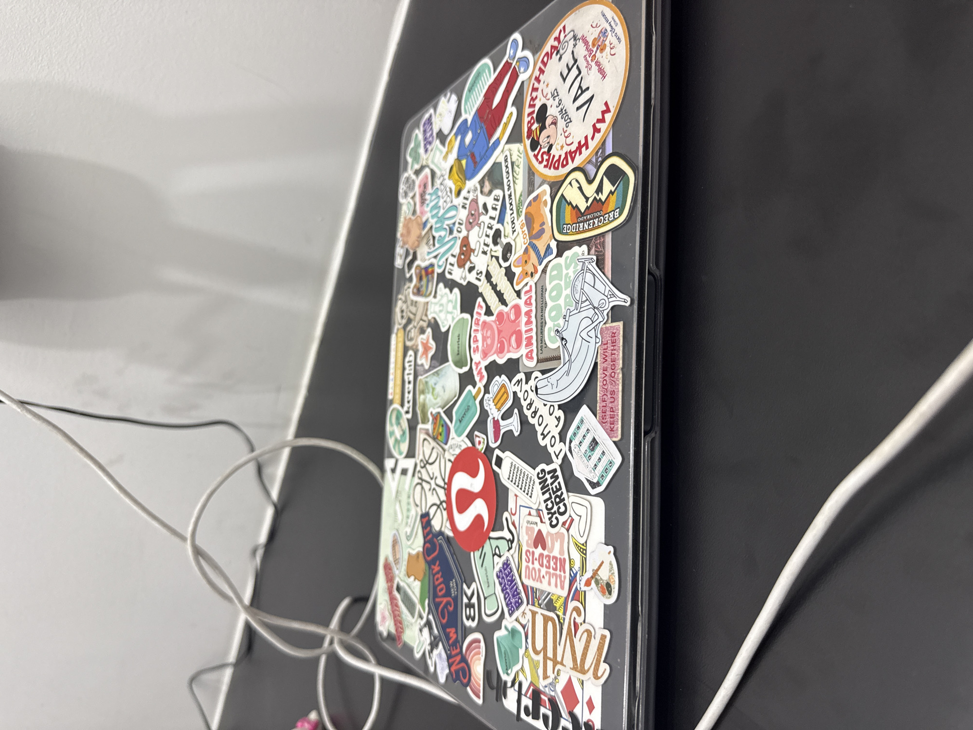 A laptop covered with numerous stickers sits on a desk with black and white surfaces. The stickers vary in design, including logos like a red "S" associated with Lululemon, text like "NEW YORK CITY", "ALL YOU NEED-IS LOVE", "CYCLING CREW", "BADASS BAKER", "MY SPIRIT", "ANIMAL", and a sticker mentioning "BRECKENRIDGE COLORADO". The sticker on the upper right corner of the laptop says "MY HAPPIEST Birthday, 2024.6.25, Happiest Birthday, TOKYO DESORT". Some stickers appear to be cartoon-themed. White cords are visible to the left and right of the laptop. The location is not discernible from the visual information provided.
