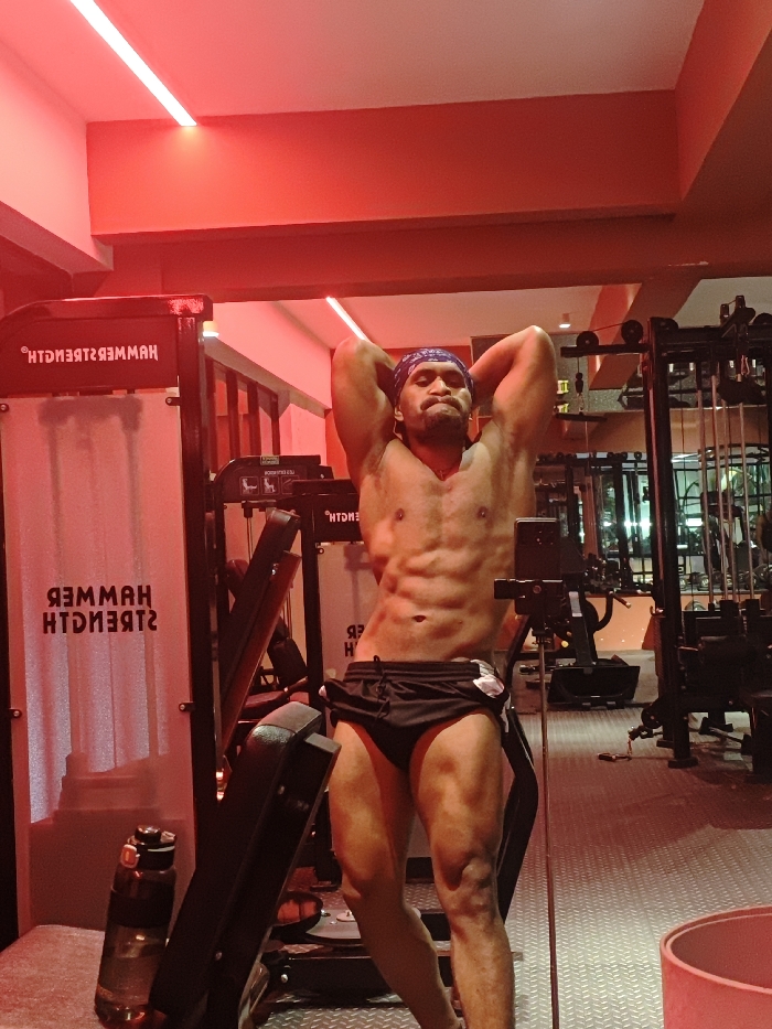 The image captures an adult male posing in a gym, specifically within the Chongqing High-tech Zone, China. The man, with a muscular build and visible abdominal definition, is shirtless, wearing a blue-patterned bandana on his head and black athletic shorts over black underwear. He poses with his hands clasped behind his head, elbows flared, and a focused expression. The gym environment is illuminated predominantly by red LED strip lights on the ceiling and walls, casting a distinct red hue across the scene. To the left, a piece of gym equipment featuring the reversed text 'HAMMER STRENGTH' is visible. A black tripod with a mounted black smartphone is positioned in the foreground, facing the man. Various other dark-colored gym machines and racks are present in the background. The floor is covered with metallic industrial grating, and a clear water bottle is visible on the floor near the left equipment.