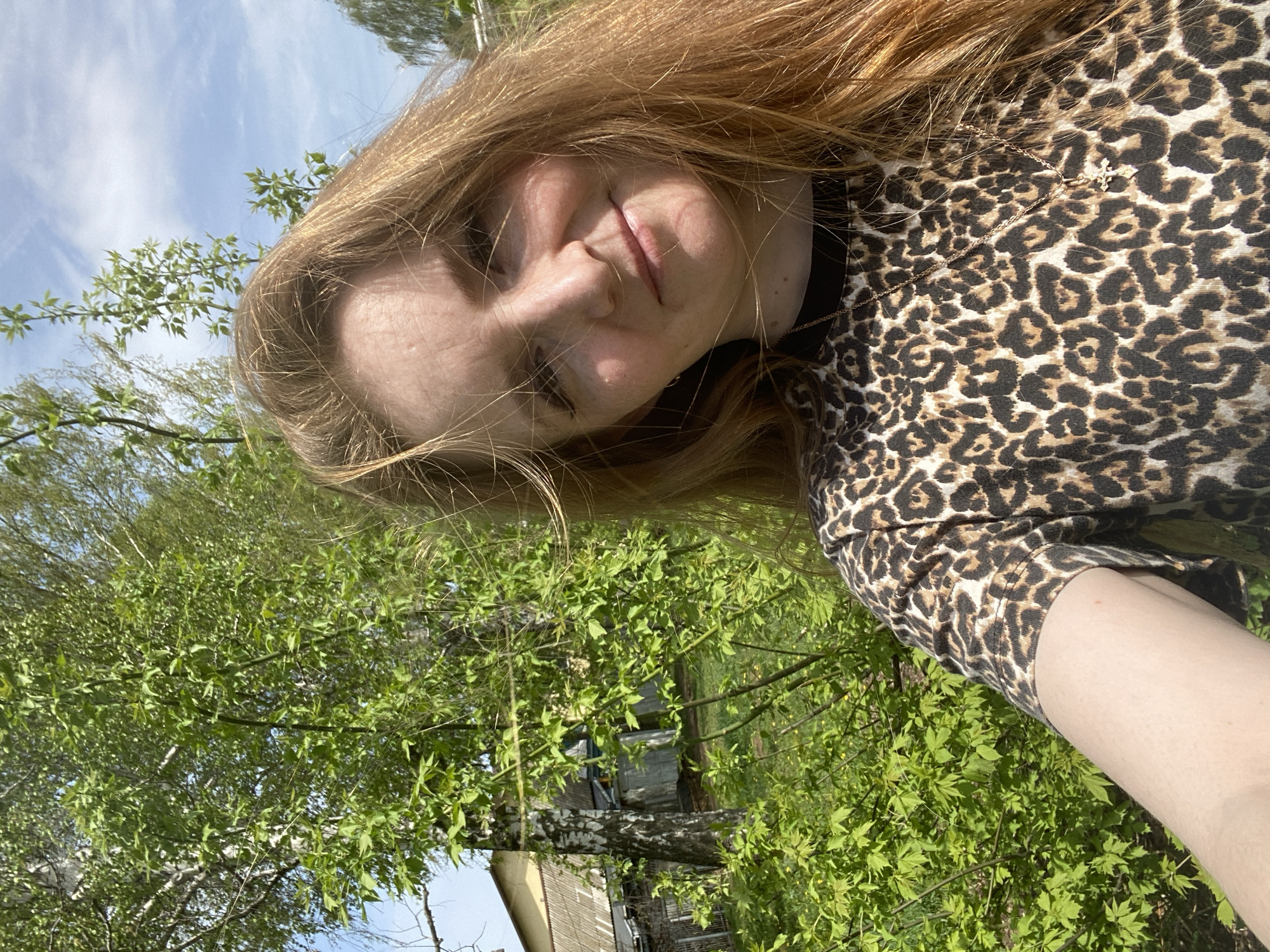 A woman with long, light brown hair is shown in a selfie against an outdoor background. She is wearing a leopard-print top and a gold necklace with a cross pendant is visible. The background consists of trees with green leaves, a building, and a blue sky with light clouds. The photograph appears to have been taken in a park or green area. The location is the city of Chaikovsky, Russia.