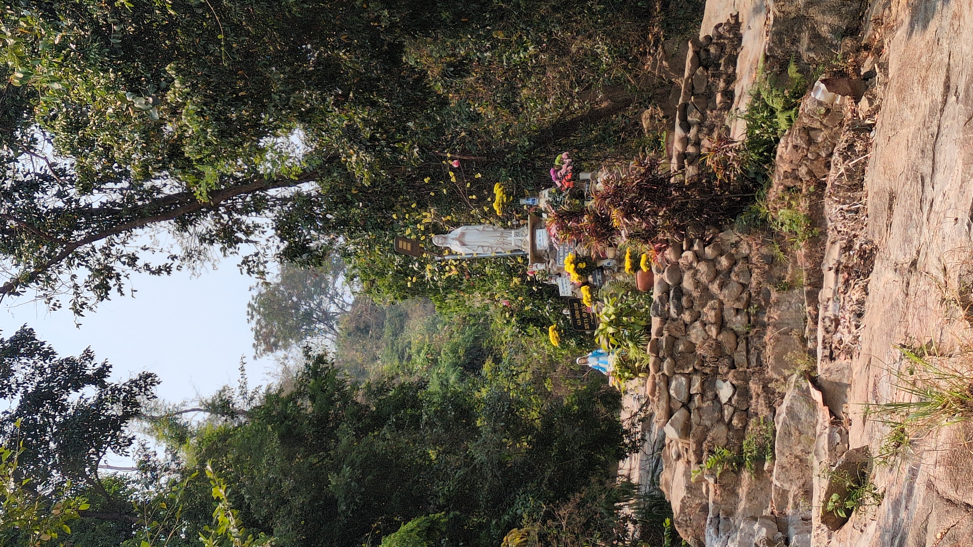 The image depicts an outdoor scene in Lâm Sơn, Vietnam, featuring a stone stairway leading up a hillside. The stairs are constructed from rough-hewn rocks and are flanked by vegetation. To the left of the stairs, there is dense foliage, including trees and shrubs. A white statue, likely religious in nature, is visible amidst the trees, adorned with flowers and signs. Along the stairway, there are additional flower arrangements and religious objects. The sky is visible in the upper left corner, and the overall impression is that of a natural, religious site in a mountainous region.