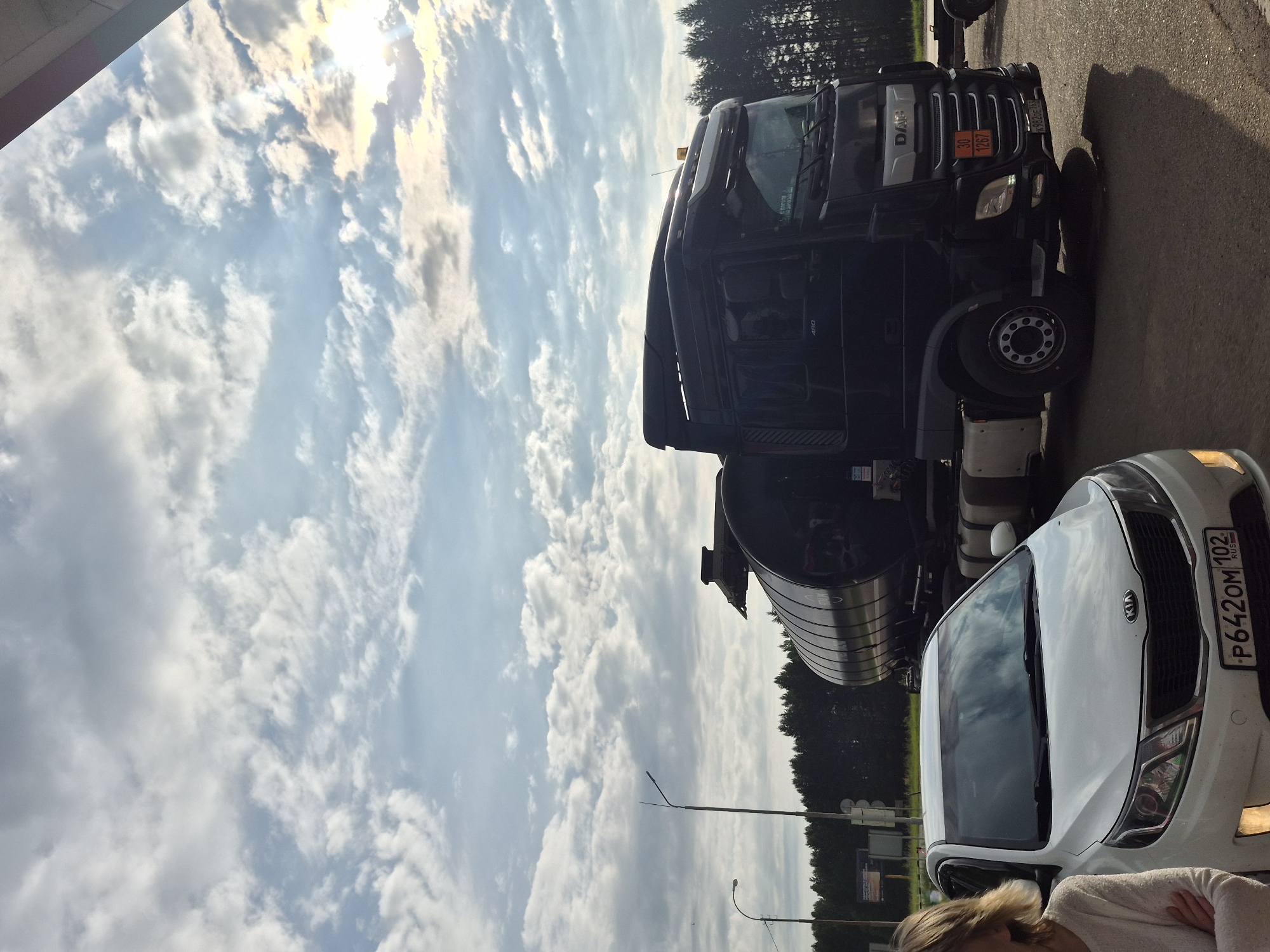 In the city of Mozhary, Russia, a black DAF tanker truck and a white Kia Rio are parked on an asphalt surface. The tanker truck is positioned to the right of the Kia, with its trailer extending behind it. A woman stands to the left of the Kia, with her arms crossed. The sky above is filled with a mix of clouds and sunlight, suggesting it is daytime. In the background, a line of trees forms a dark silhouette against the horizon. The scene appears to be a roadside or parking area.