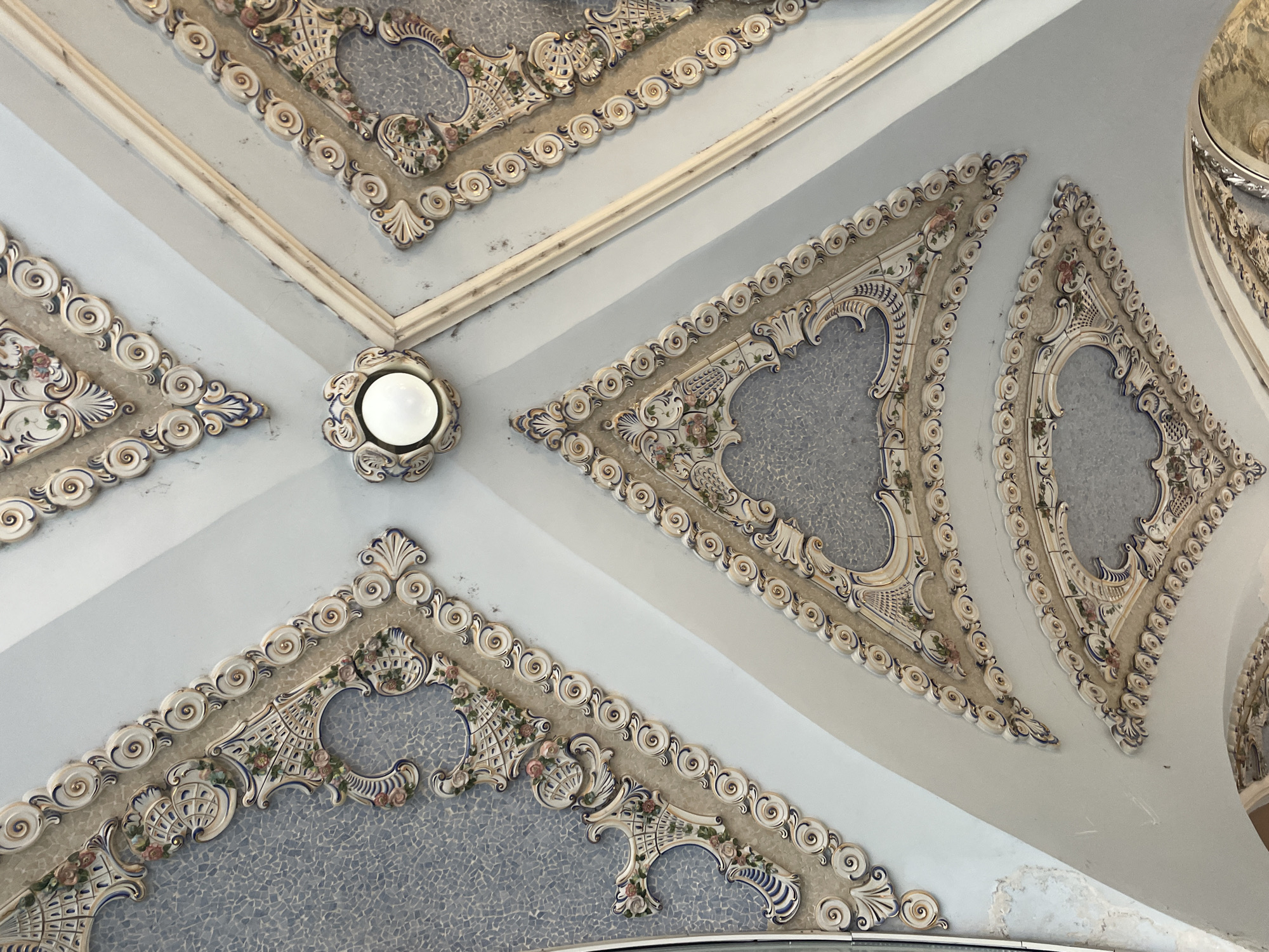 This is a close-up, low-angle shot of an ornately decorated ceiling in a building, possibly a historic or public space. The ceiling is divided into triangular sections with a light blue base color. Each triangle is bordered by elaborate plasterwork or molding featuring intricate designs of swirling patterns, floral motifs (possibly roses), and geometric shapes, all in a muted palette of white, beige, and touches of gold and blue. The centers of the triangular sections appear to be filled with mosaic tiling. A single light fixture with a bulb is situated at the intersection of several triangular sections. The overall style suggests an Art Nouveau or Belle Époque influence. This image likely originates from Batumi, Georgia, given the context of previous queries.